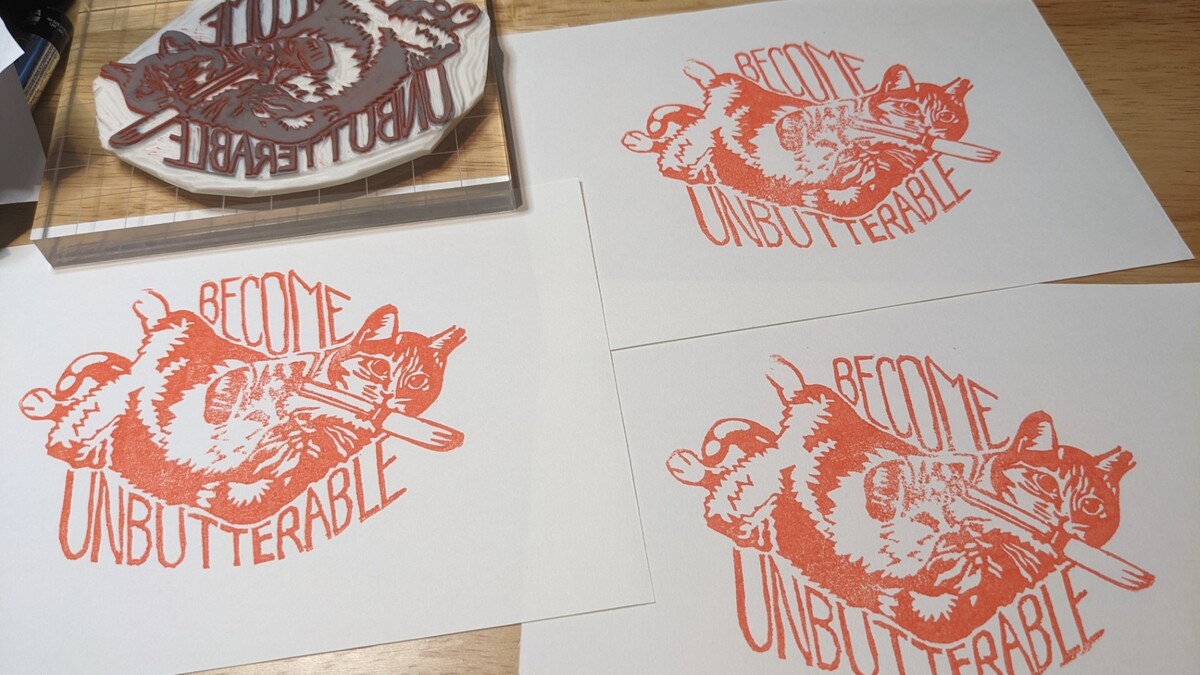 3 copies of the same stamp in orange ink are spread out next to the hand carved rubber stamp they were made from. They show a cat lying on his back with paws curled, holding a butter knife in his mouth. Text around the cat reads, in all caps, 'become unbutterable.'