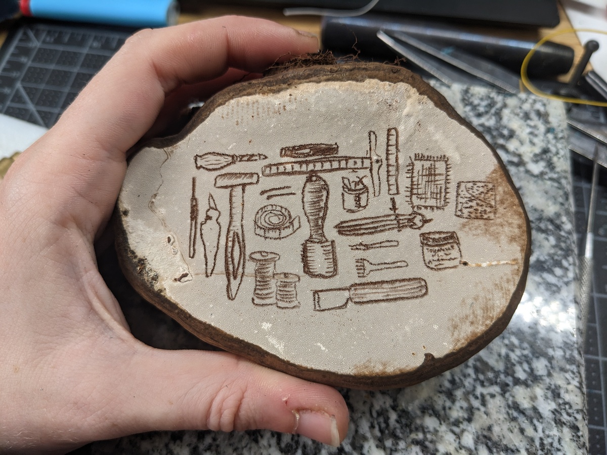 A hand holding a mushroom with a whitish surface. Scratched into the surface and showing up as brown lines is an illustration of many leatherworking tools.