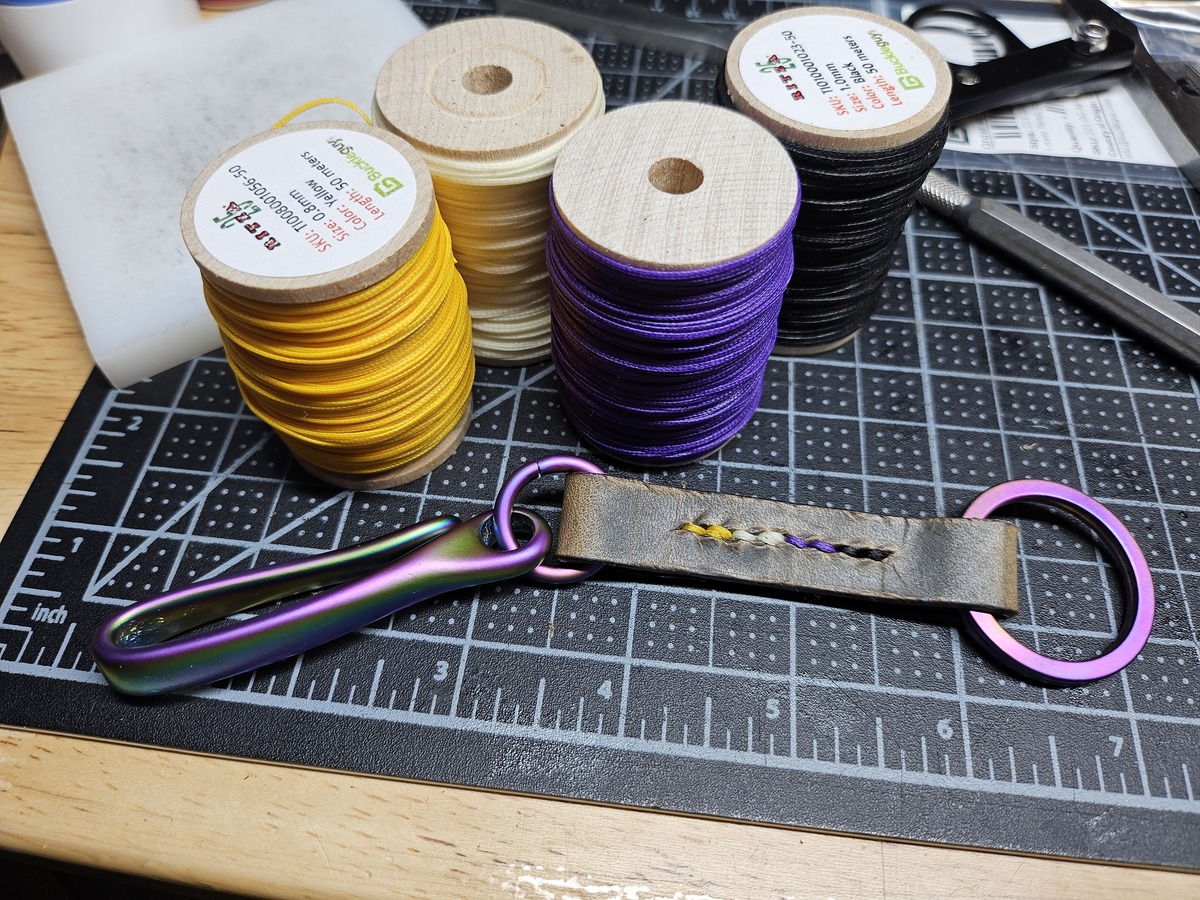 a keychain with an iridescent fishhook style attachment linked via leather to an iridescent keyring. the leather is stitched with nonbinary flag colors.