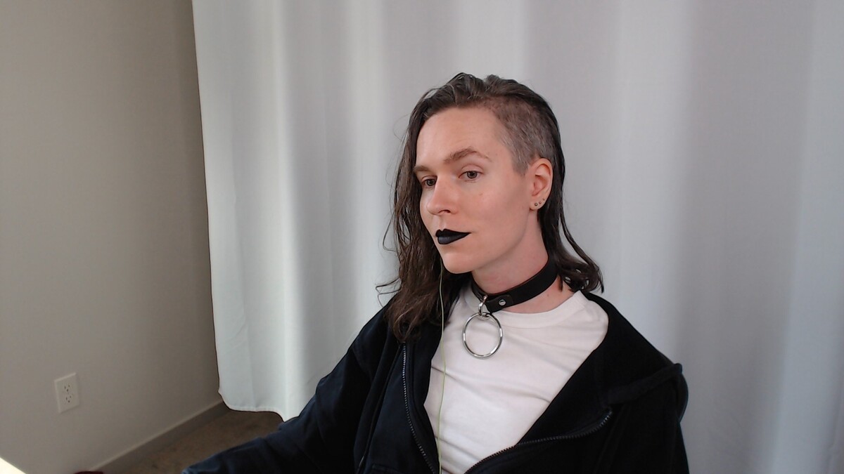 A white woman with a shaved side cut wearing a black leather collar with a large dangling o-ring.