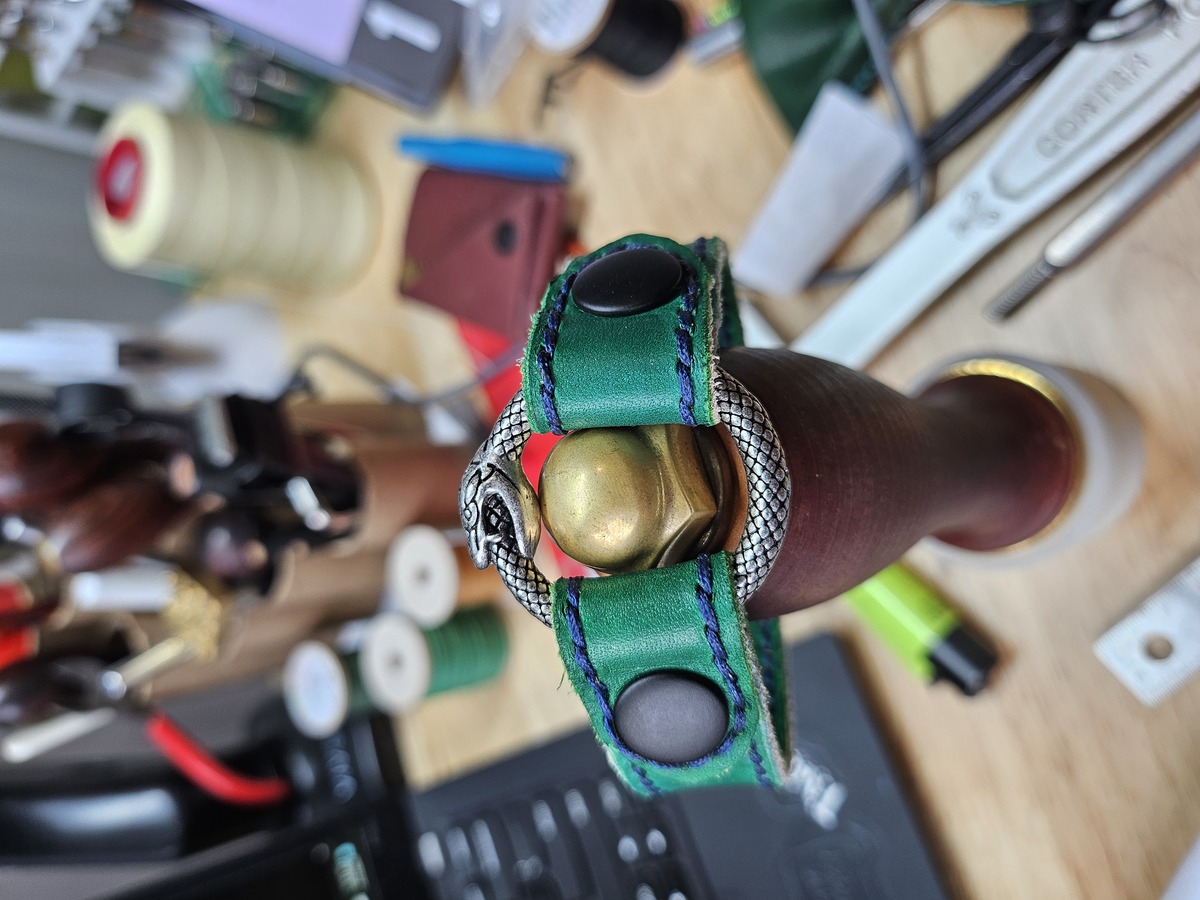 A green leather bracelet, stitched along the edges with dark blue thread, holds an ouroborous o-ring in place with two black snaps.