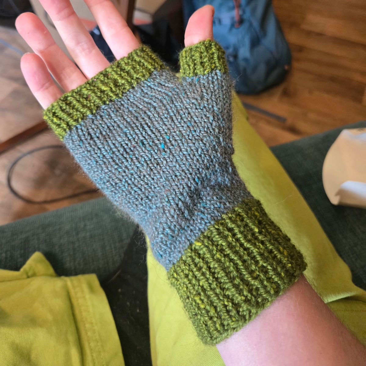 a hand wearing a knitted fingerless mitten. it's knit in a slubby, almost tweedy yarn, with the body being blue grey stockinette and the cuffs and tips a vibrant green rib.