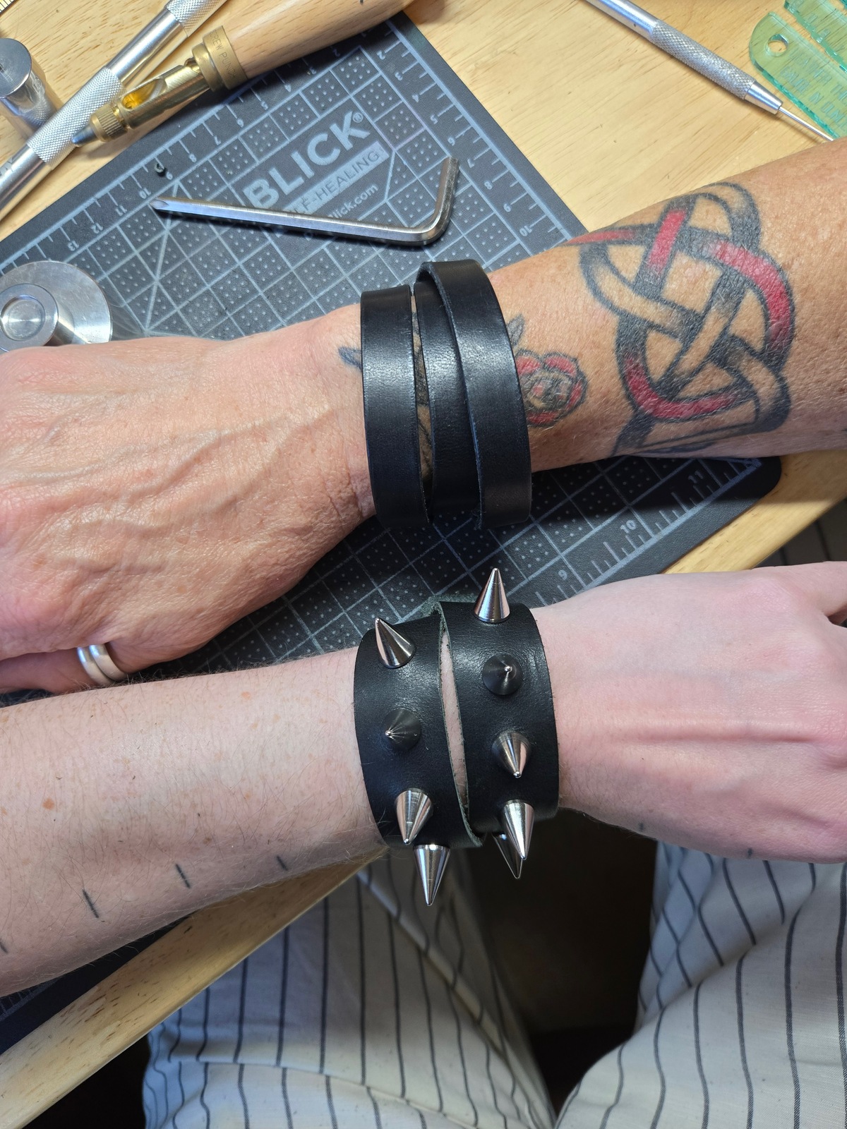 two wrists, each wearing a black leather wrap bracelet. the upper bracelet is a thin strap wrapped 3 times around the wrist. the lower bracelet wraps twice, with a thicker strap, and has carefully placed spikes that avoid the wrap spots.