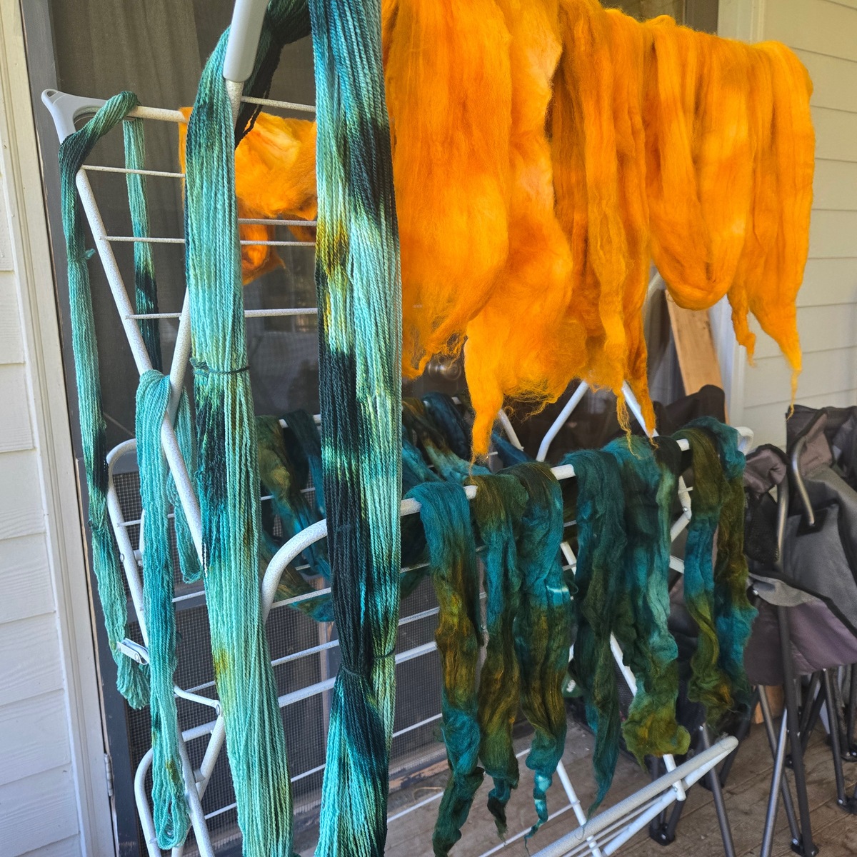 4oz of yarn and a pound of wool, chunked out, drying on a drying rack outdoors. the yarn and half the wool is a mix of teals and greens; the other half of the wool is a beautiful orange-gold.