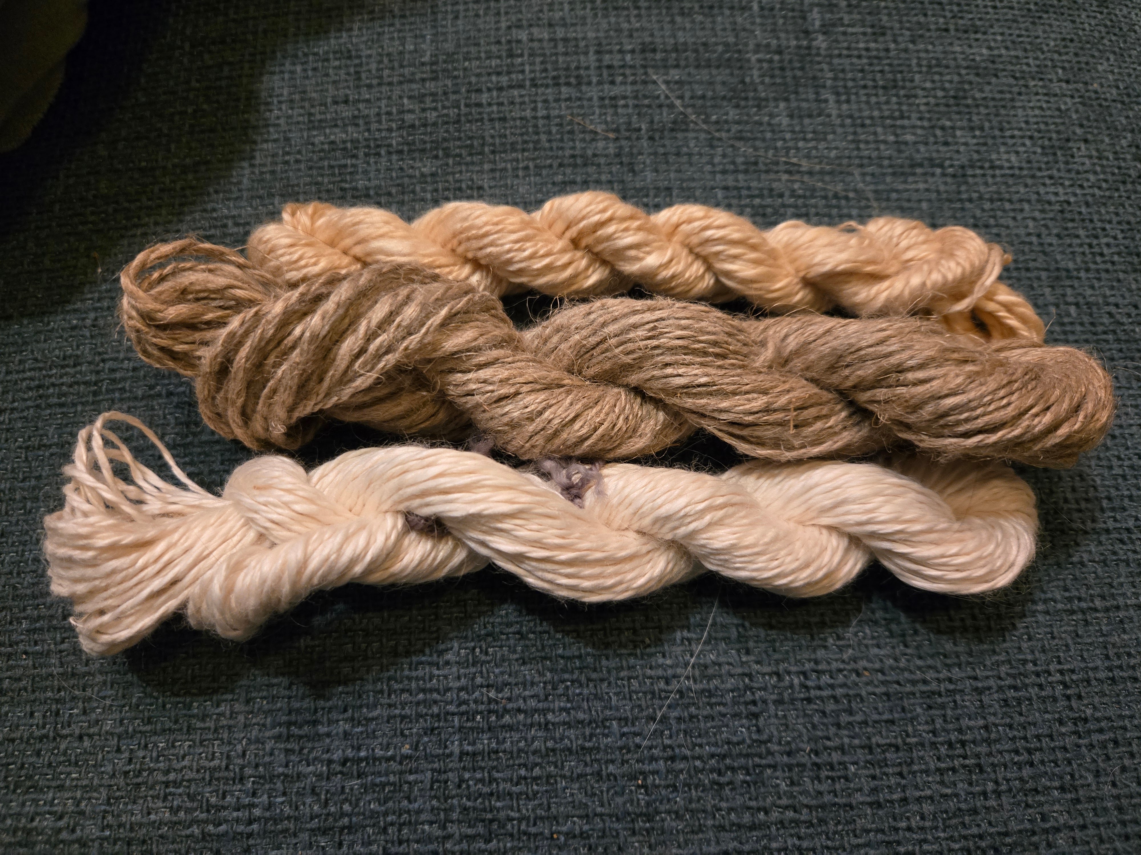 3 small skeins of handspun yarn, each made from a different plant fiber. the top skein is probably, though not definitely, lotus, and it has a lovely golden color and looks shiny and soft. the middle skein is flax, which looks quite rough and is a medium brown. the bottom skein is banana fiber and is white and shiny