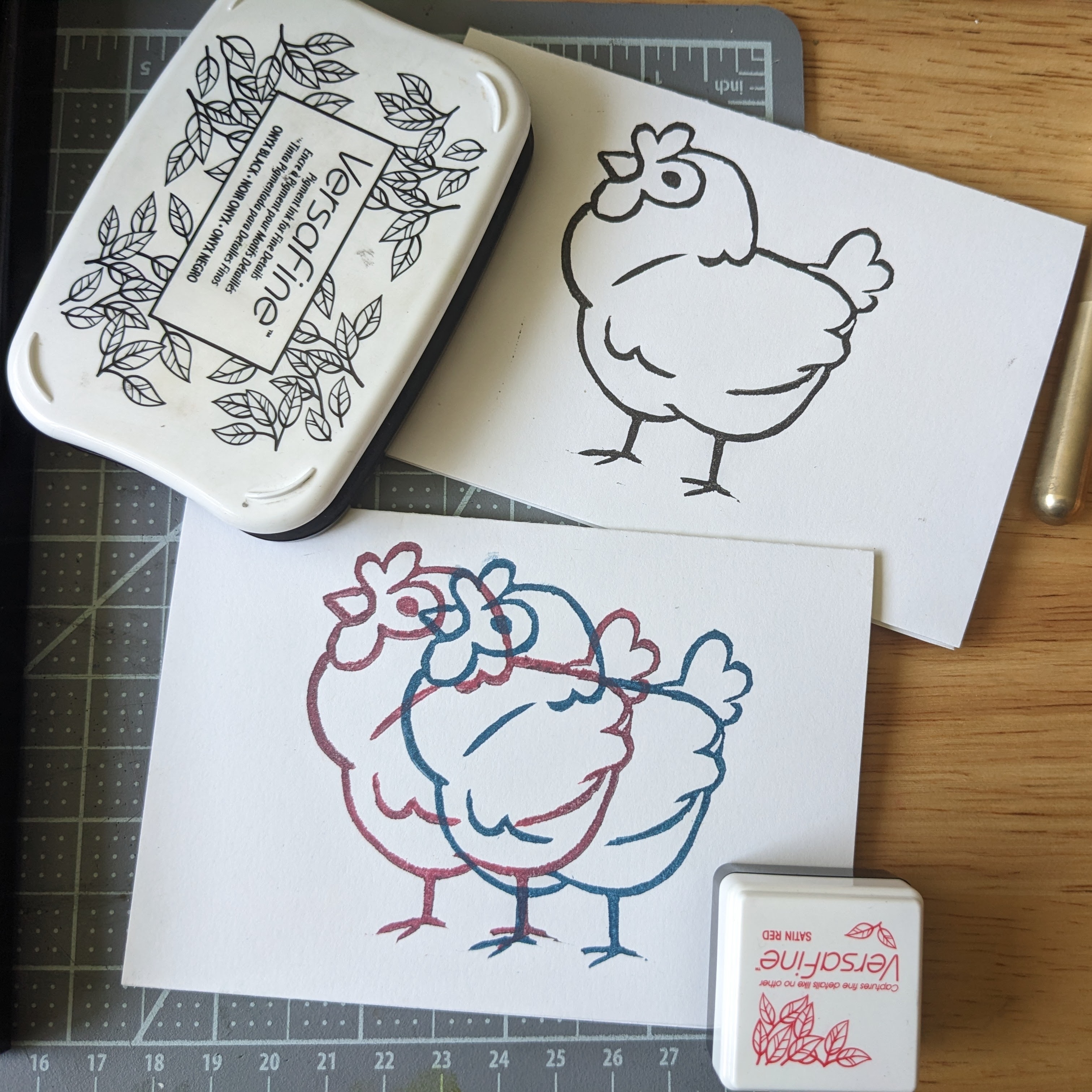 2 white greeting cards with the Artisans Cooperative logo, a chicken. One card has a single print of the chicken in black ink, and the other has two overlapping prints in blue and red ink