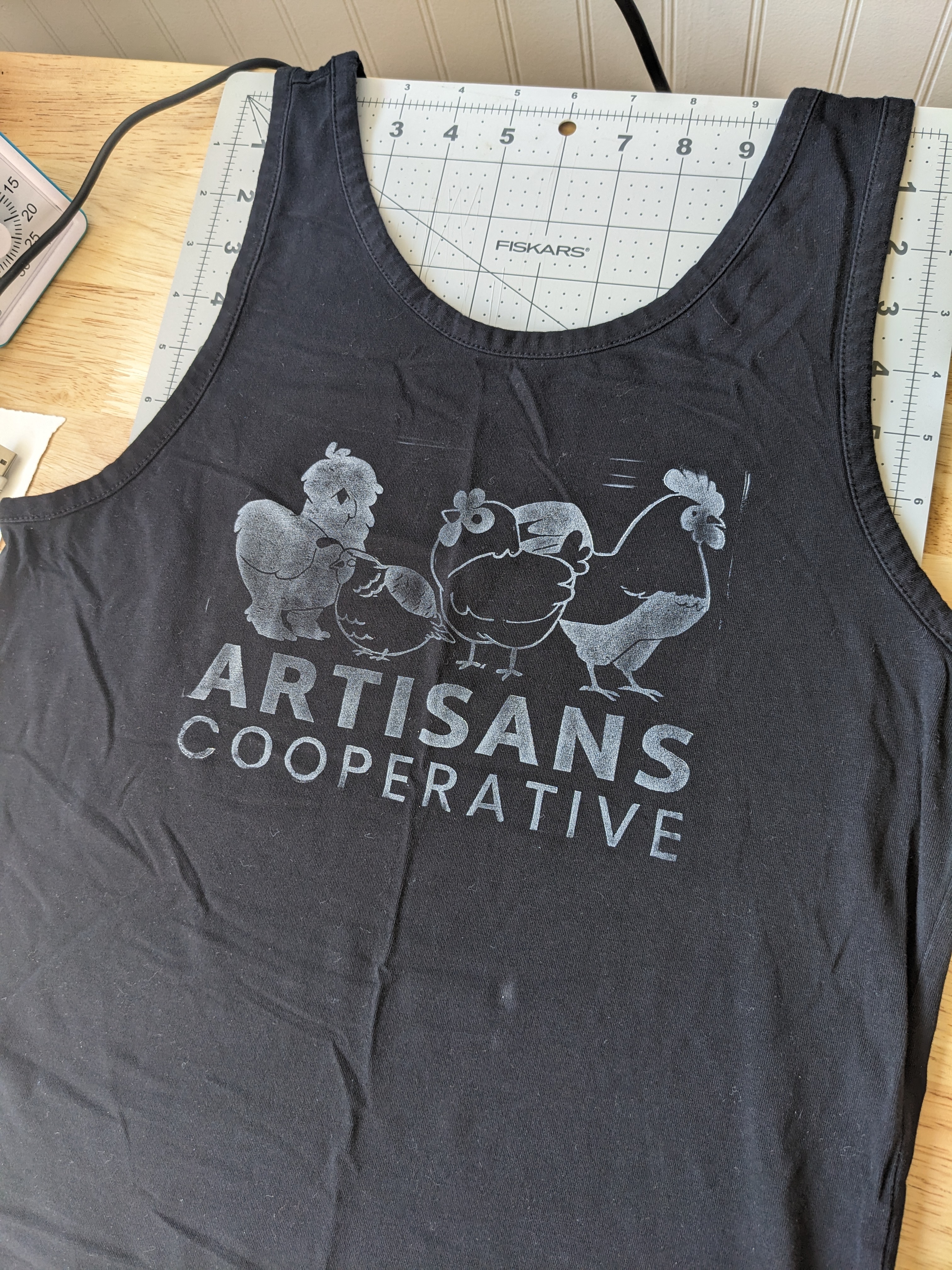 A black tank top laid on a desk. In white ink it reads 'Artisans Cooperative' with a print of some chickens and a quail.