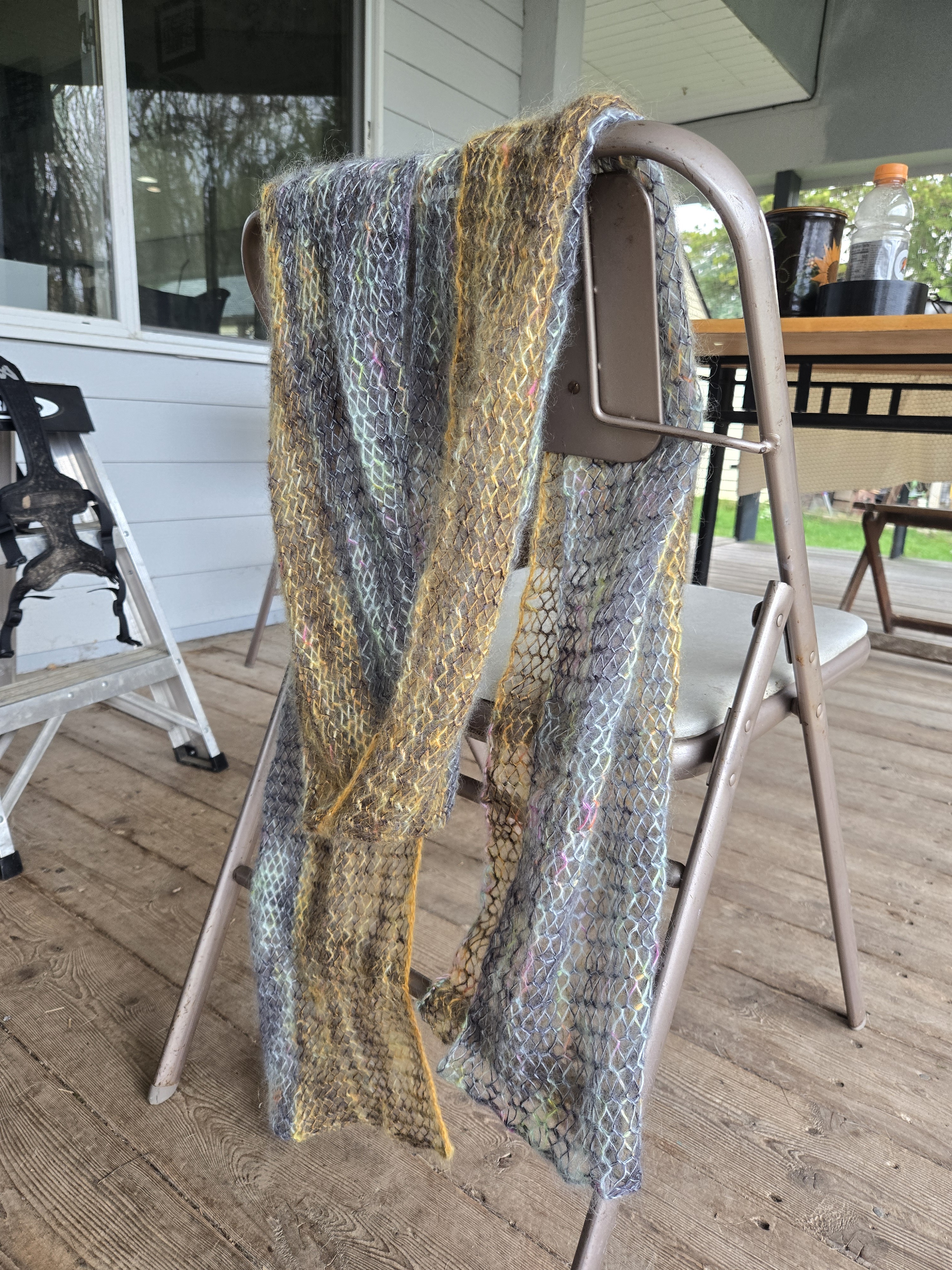A diaphanous knit lacework scarf draped over the back of a chair. It is split down the long way into two colors - one tinted orange and one tinted mint blue. Both colors, the orange and the blue, are held double with the same variegated gray, making the piece more cohesive. The yarn overs in the lacework create airy repeating holes.