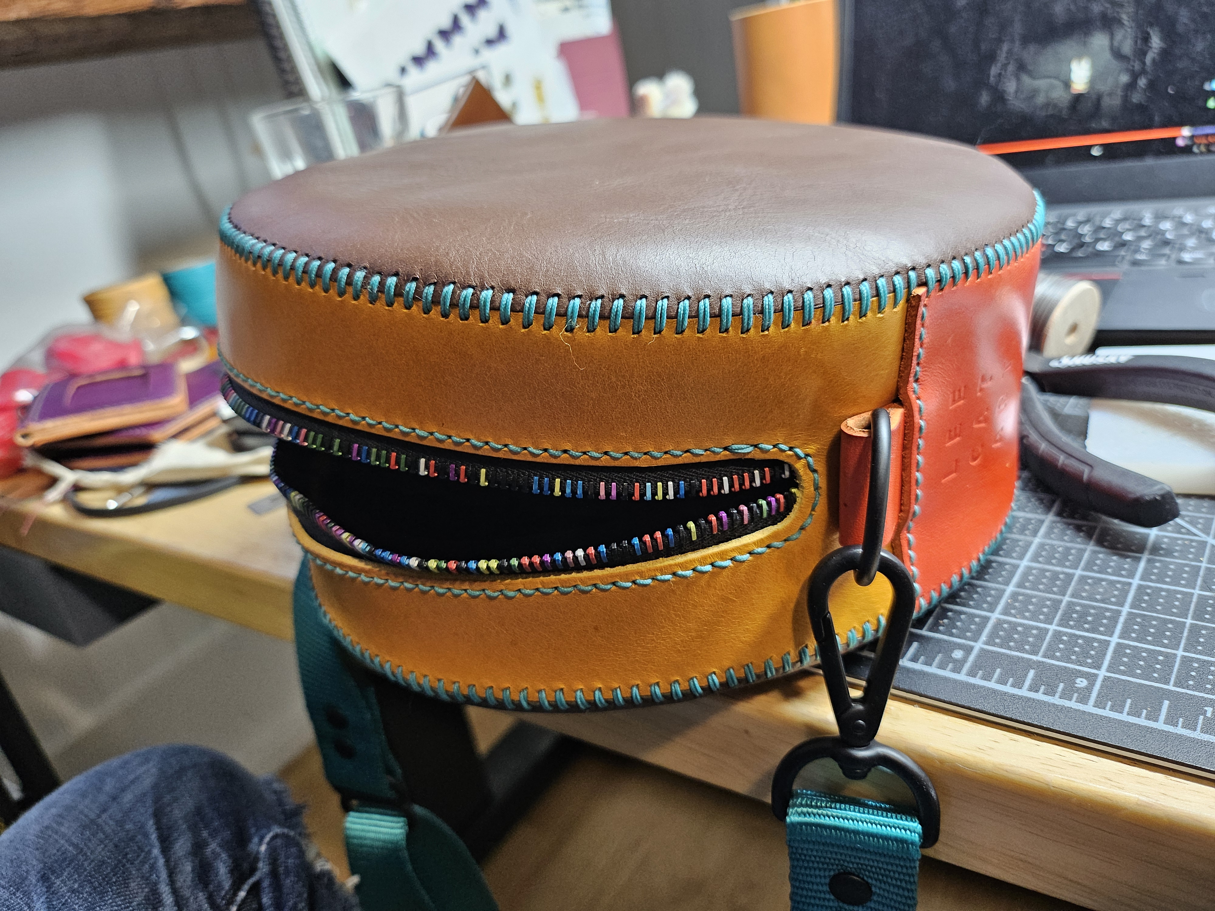 A round bag in brown, mustard yellow, and rich deep orange, with a teal shoulder strap.