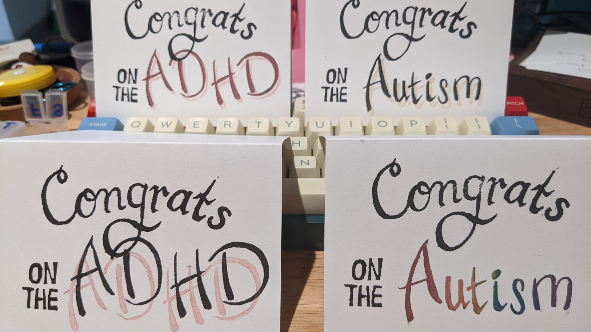 4 greeting cards propped up on a keyboard. On the right hand side, two cards read 'Congrats on the Autism'; one in rainbow ink and one in black ink with a glittery gold shadow. On the left, two cards read 'Congrats on the ADHD'; one in red and one in black, both with glittery pink shadows.