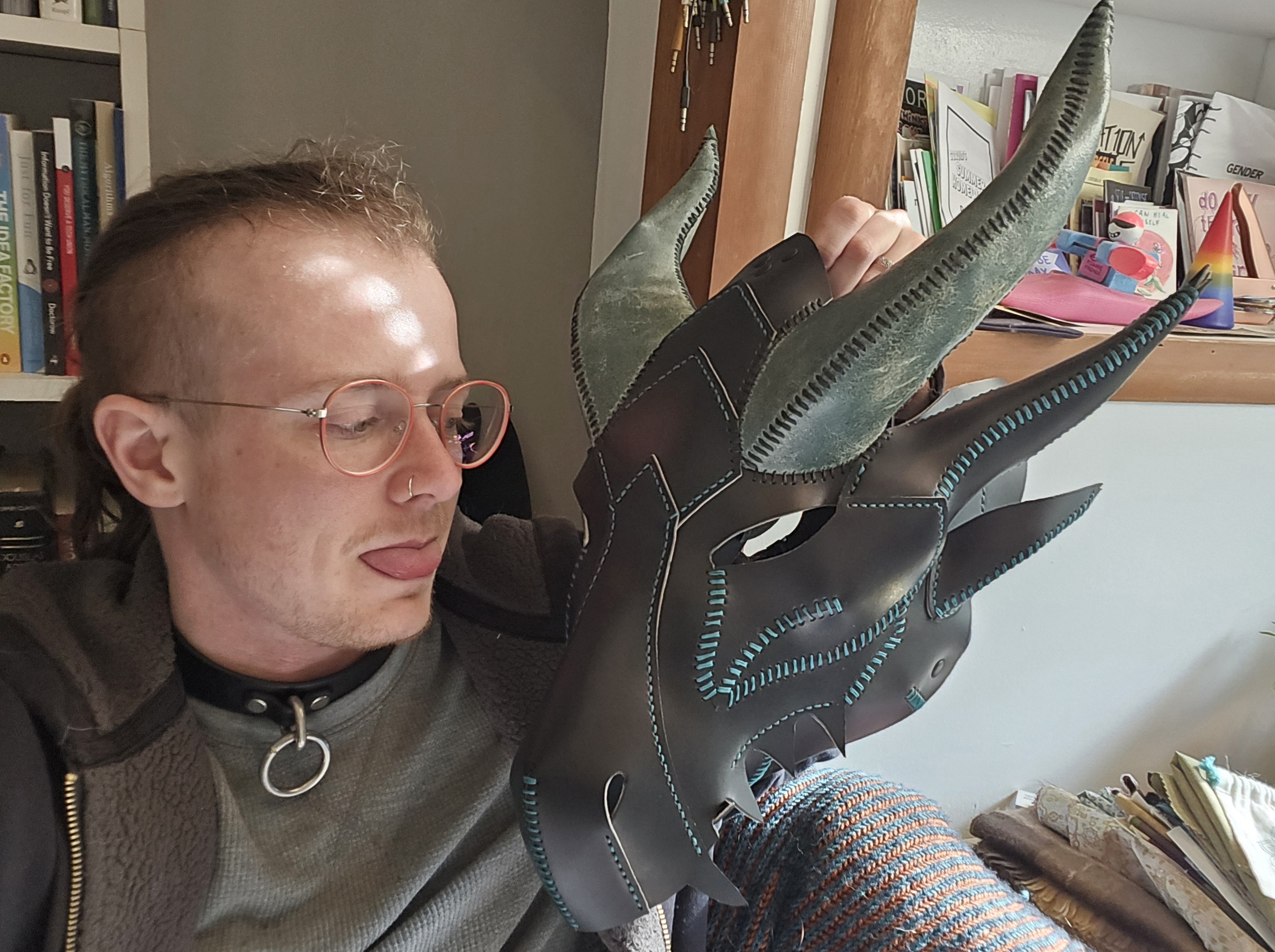 lee (a white person with glasses and a side shave) holds up a leather dragon mask in black and dark green. ze sticks hir tongue out at it.