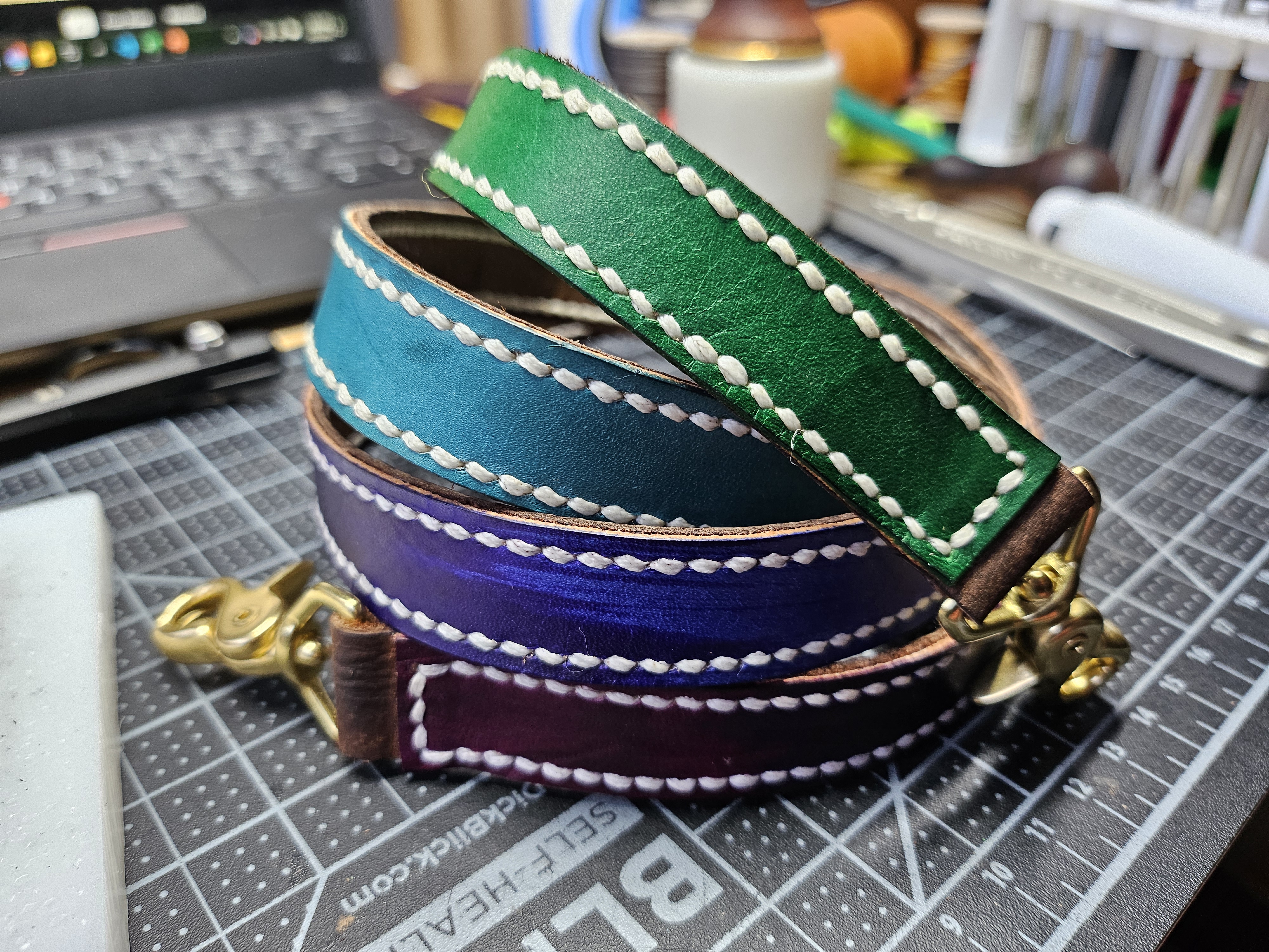 a coiled up purse strap in gradient cool colors - we can see green, teal, indigo, and a slightly pinkish purple. It has brass hardware and is stiched along its length with cream stitches.