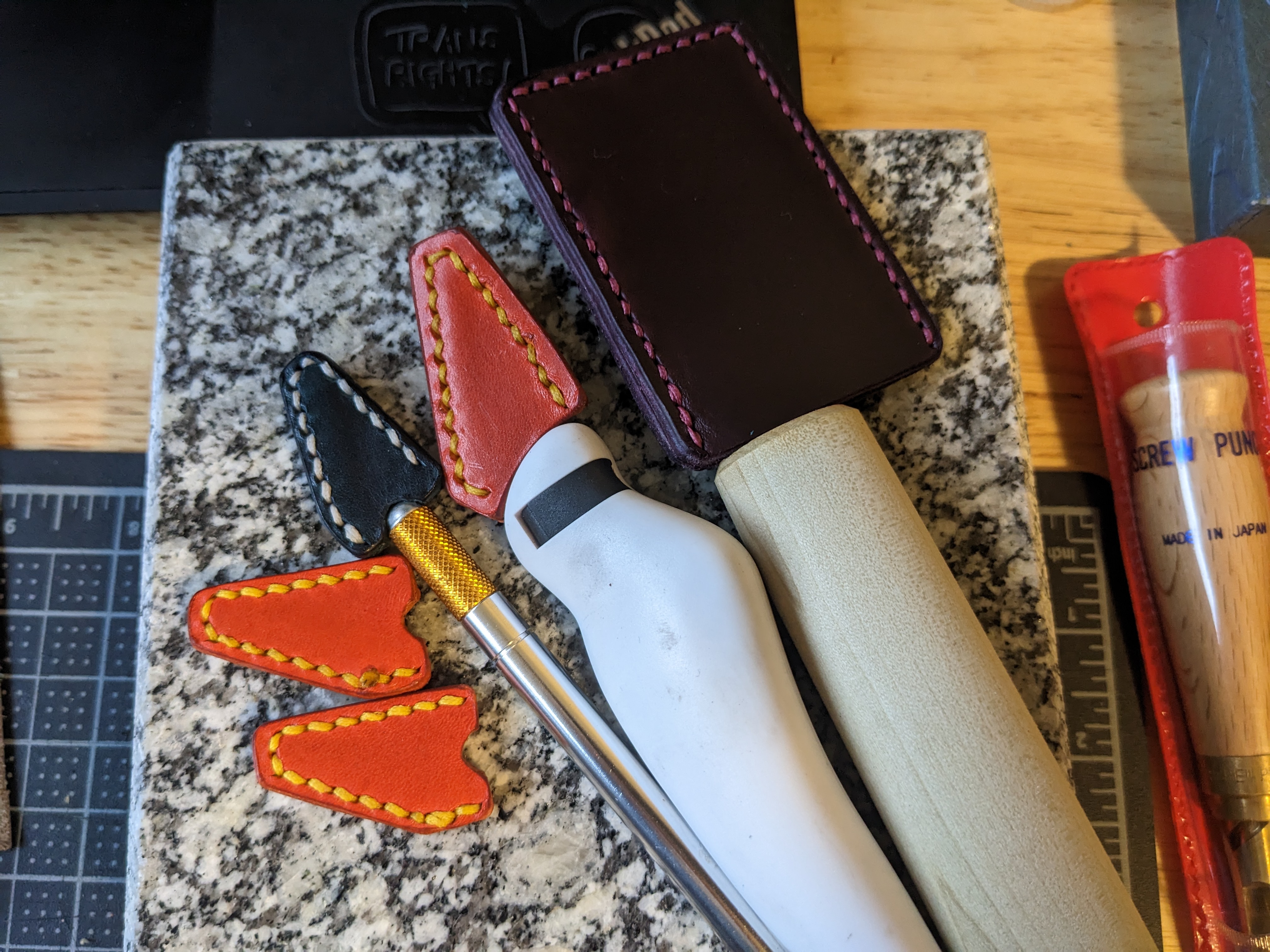 Several blades with leather sheaths, and a few extra sheaths. There's a #2 blade with an orange sheath with yellow stitching, a #11 blade with a blue sheath with light grey stitching, and a skiving knife with a plum sheath and pink stitching.