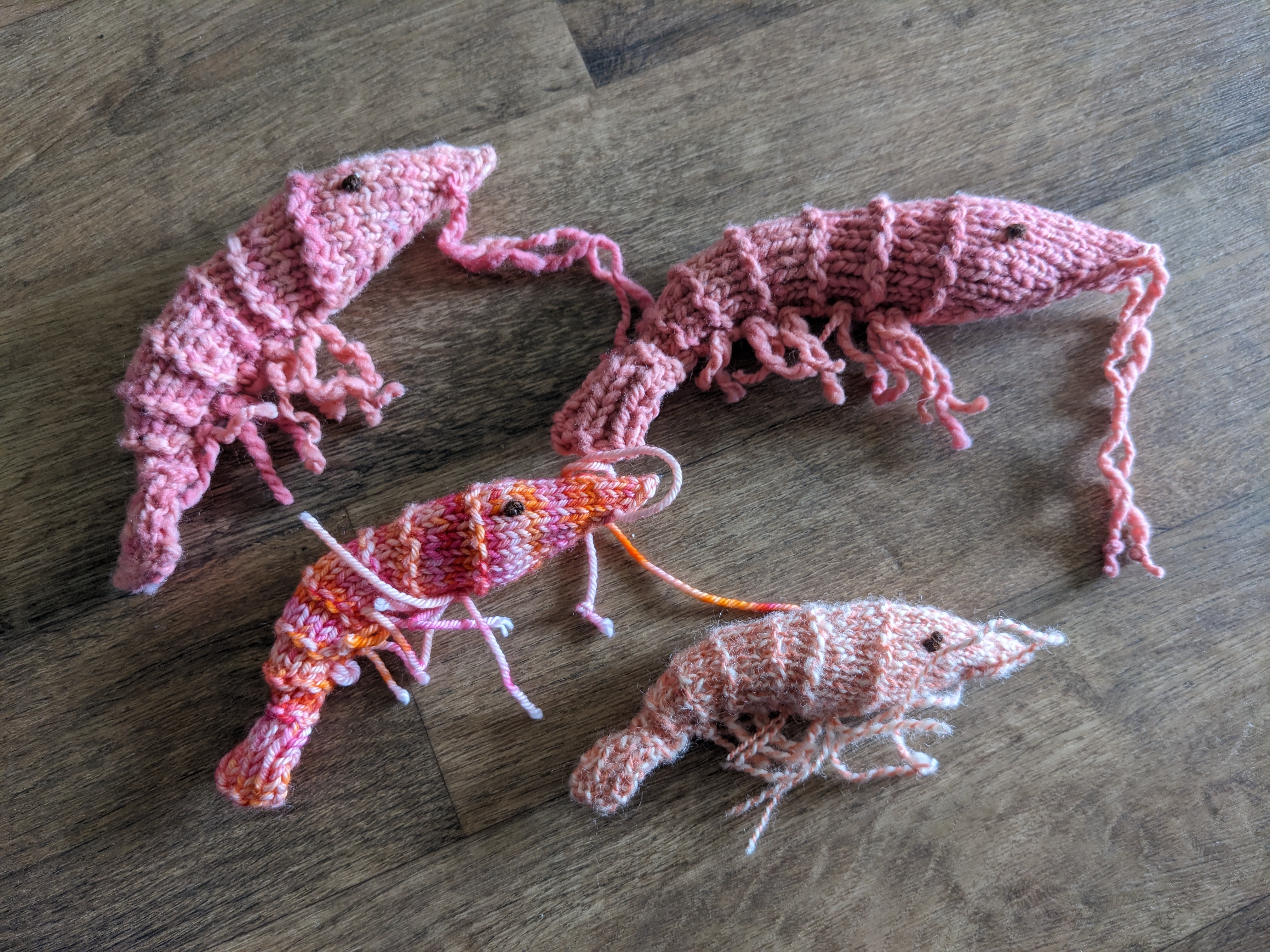 Four knitted shrimp in various shades of pink. They are all somewhere around hand-sized, and have little yarn antennae and leggies.