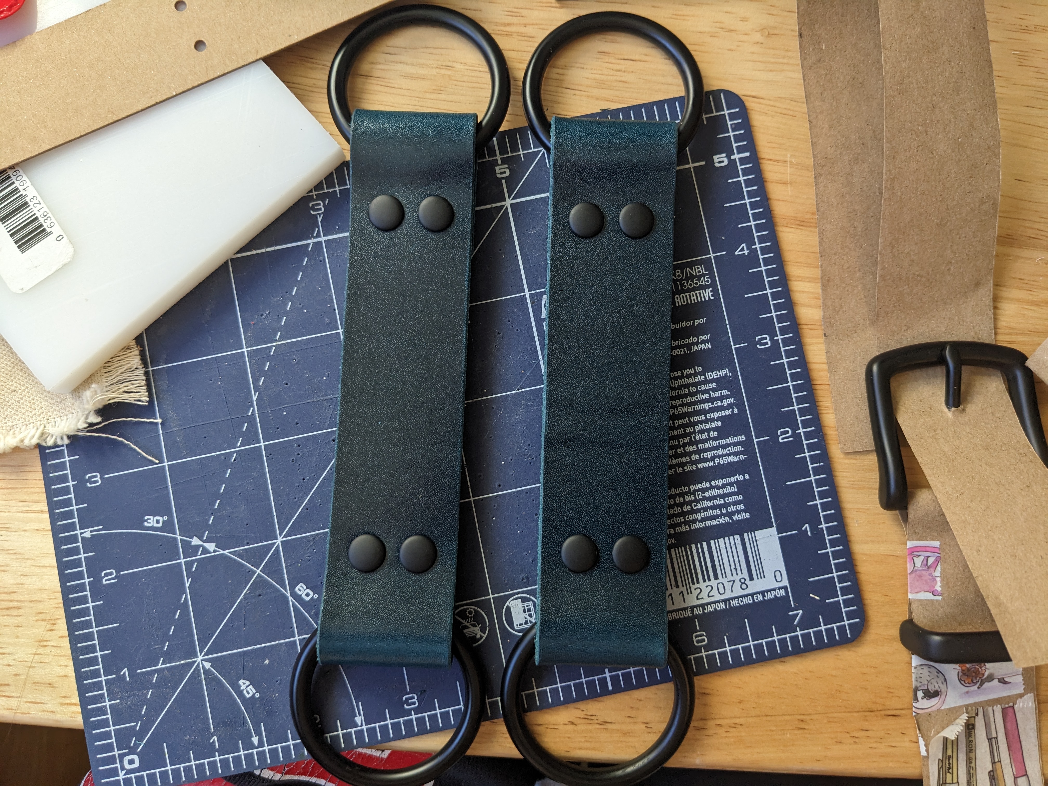 two pieces of a chest harness sitting on a messy workbench. both pieces are about 8 inches long total and consist of two large o-rings joined by a dark teal leather strap. the o rings and rivets are matte black.