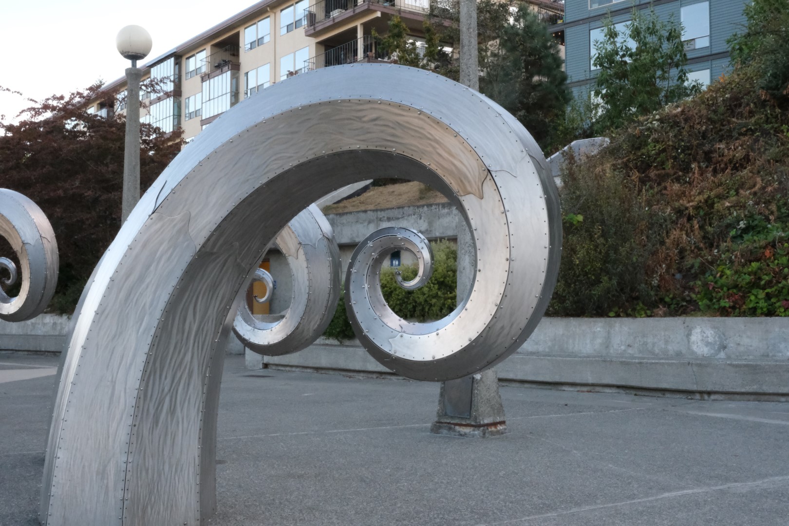Photo unrelated to content of post. A metal sculpture rises up and curls into itself, with similar sculptures around it. Looking through the very center of the curve, an any-gender bathroom sign is visible.