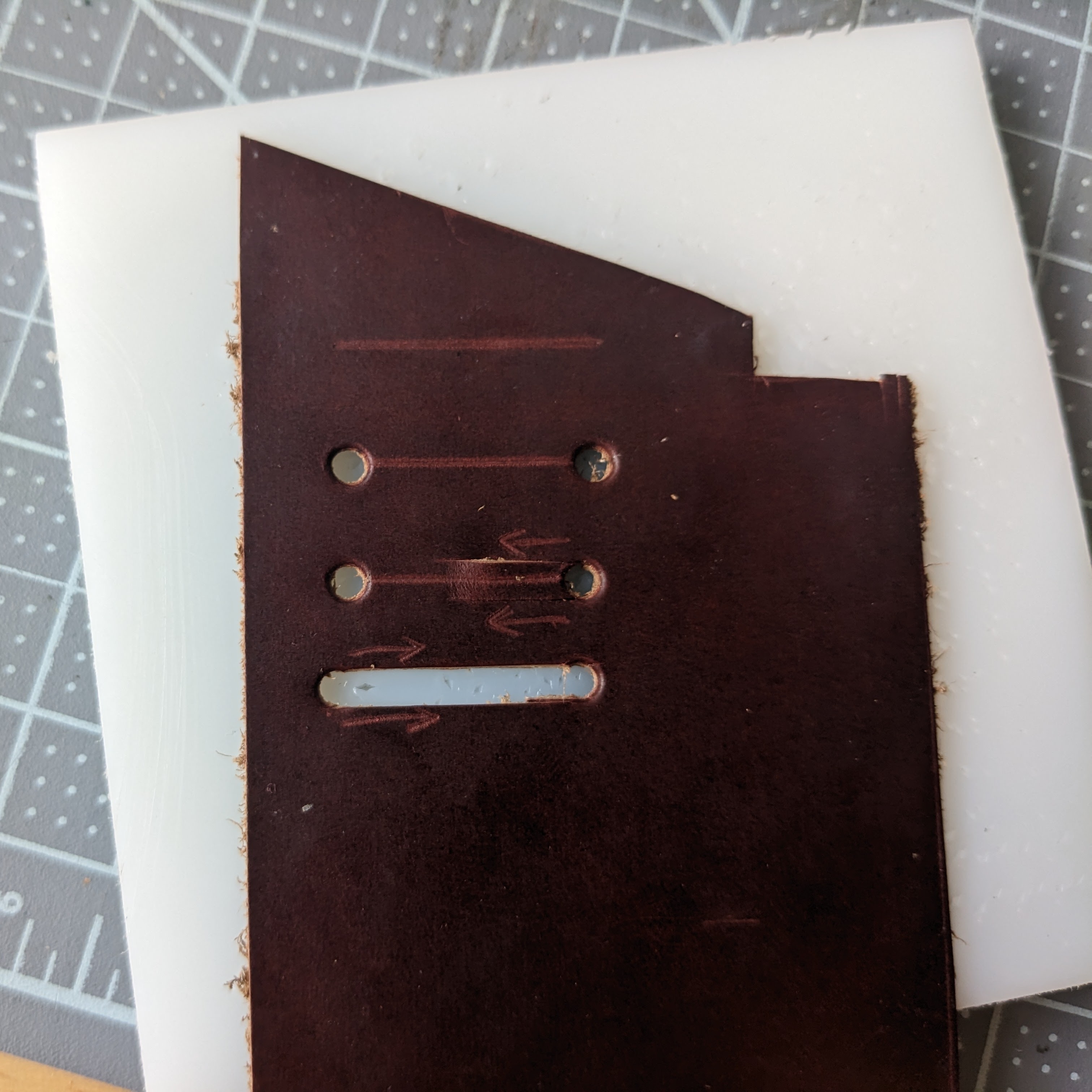 A small piece of leather showing the steps to creating an oblong hole. First there is just a straight 1-inch line drawn by scratch awl. Holes are punched at both ends of that line. Two cuts are made coming from the right-side hole inwards (shown by arrows), about halfway. Similar cuts are then made from the left-hand hole inwards to meet them.