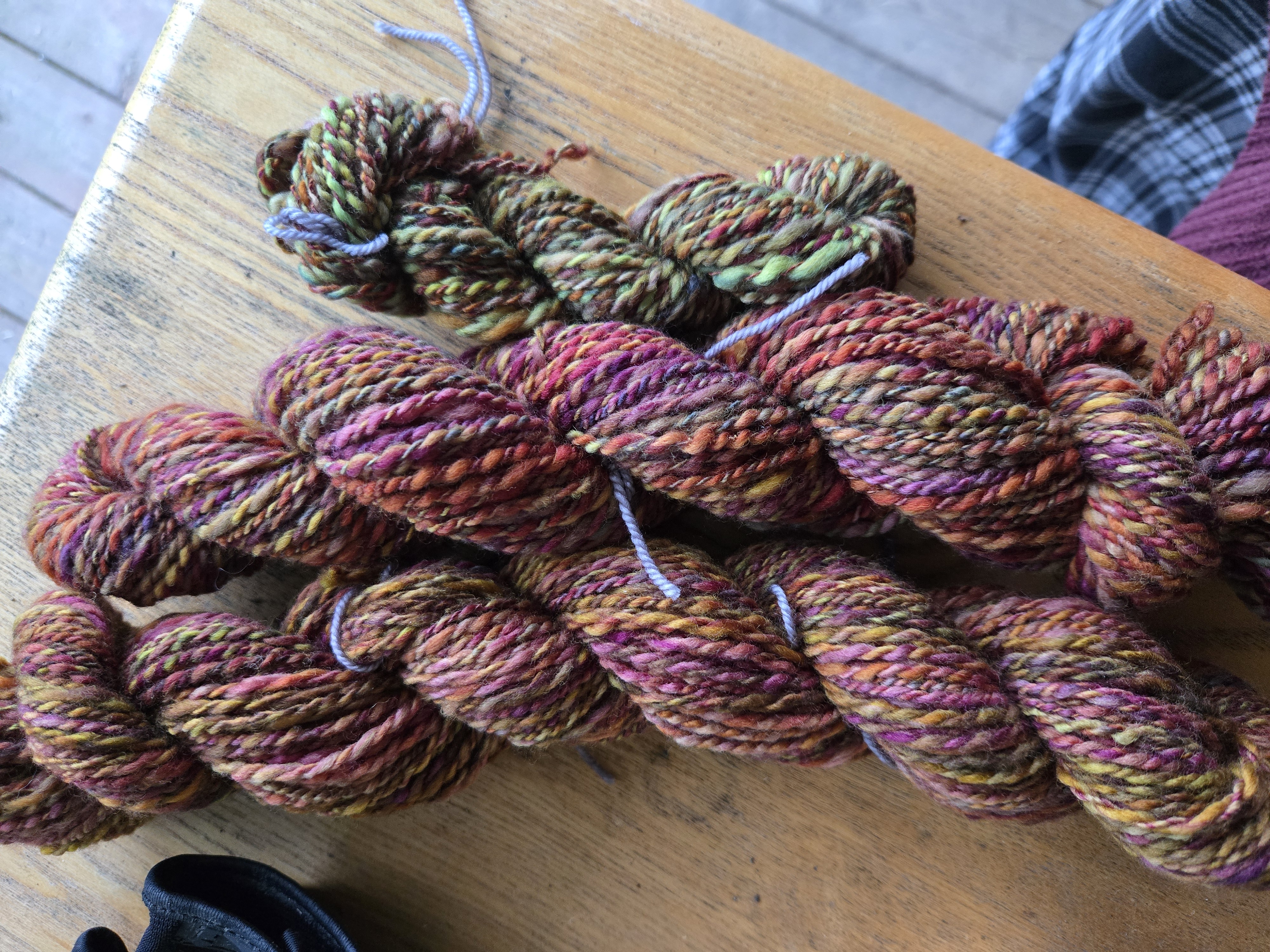 3 skeins of handspun yarn, 2 large and 1 small. the large ones are a rich earth-tone blend of reds, pinks, browns, and hints of green and gold. the smaller skein is similar but with a decidedly greener hue