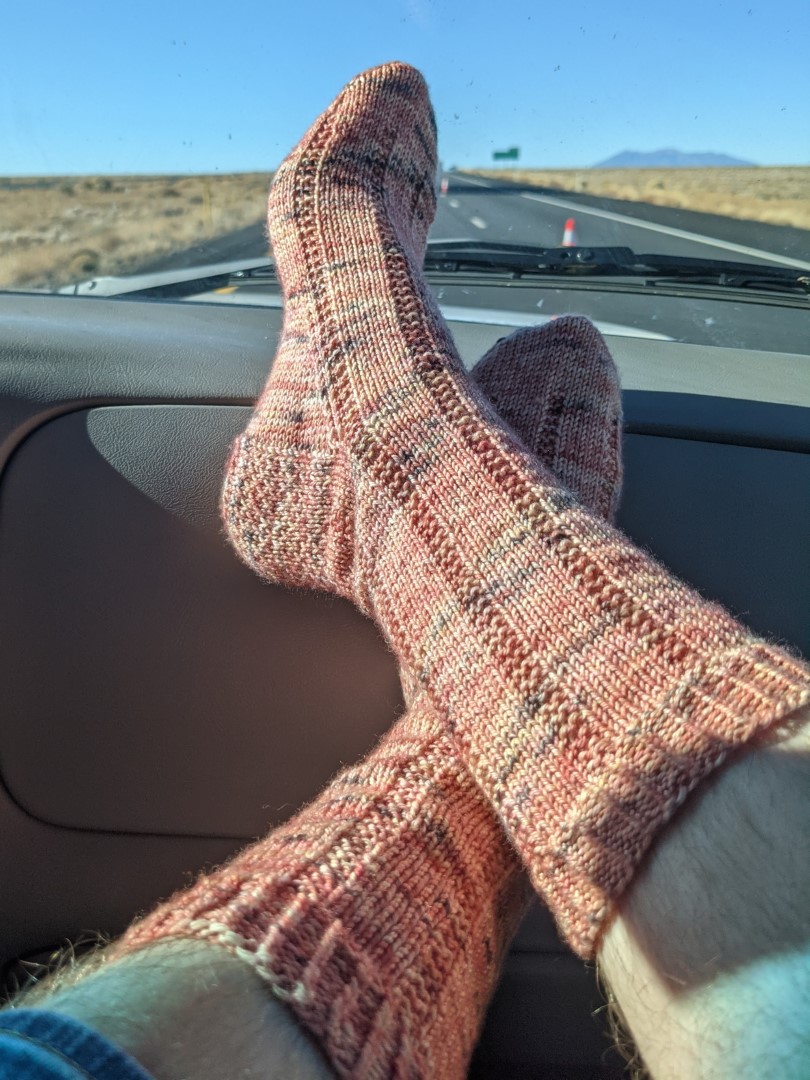 Feet propped up on a car dashboard, with a desert landscape beyond. The feet are in salmon-colored socks with black flecks, and decorative lines running down the socks.