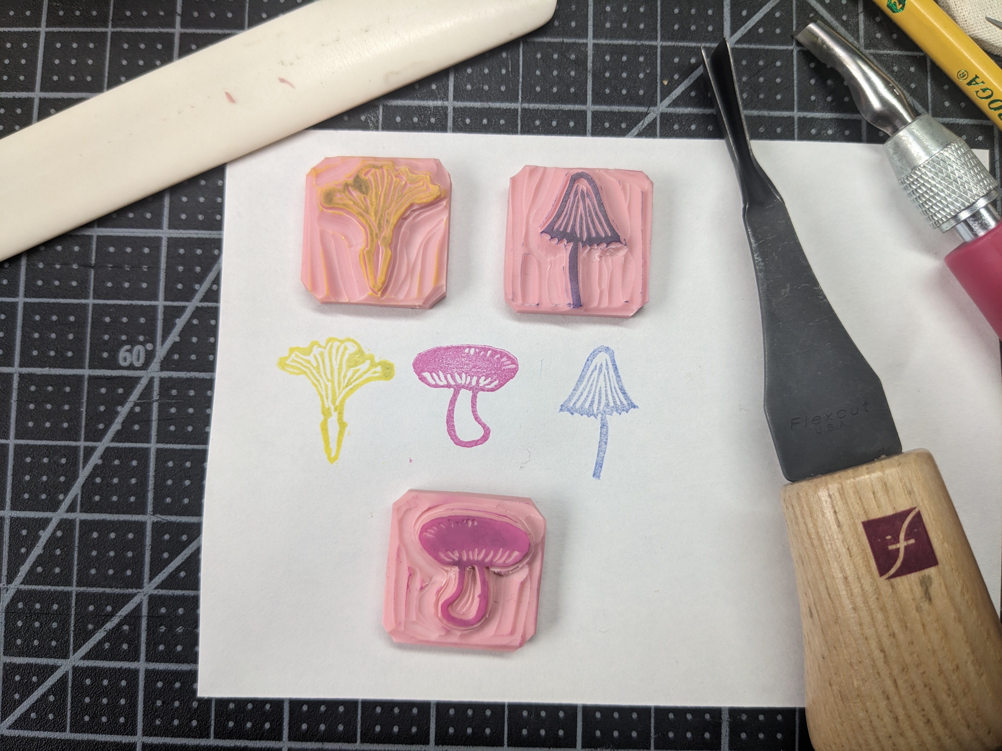 3 tiny mushroom stamps next to their impressions. They are all about 1 inch square. There is a chanterelle in yellow, a russula in pink, and witch's hat mycena in indigo.