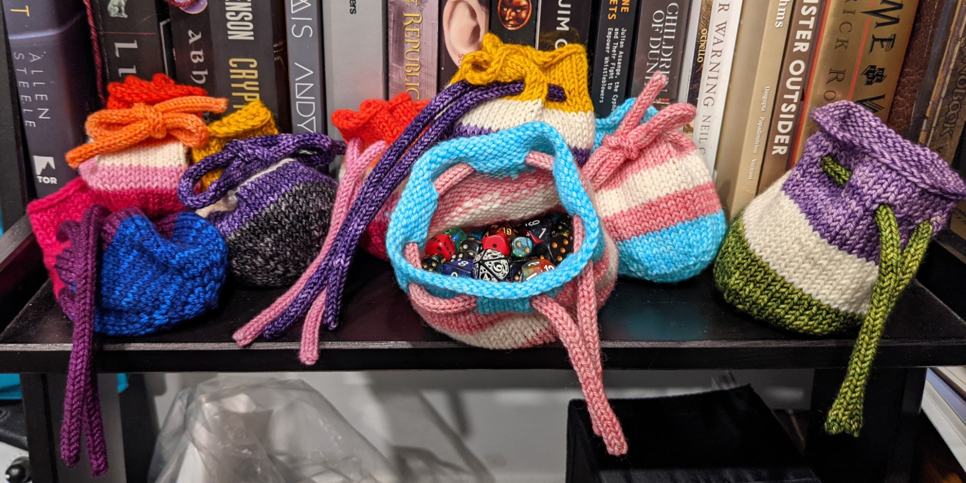Several knitted drawstring dice bags sit in front of a bookshelf. They are in different pride flag colors; from right to left (skipping a few duplicates) bisexual, lesbian, nonbinary, trans, and genderqueer. The trans-colored dice bag in the center opens towards the camera, showing a variety of colorful dice inside.