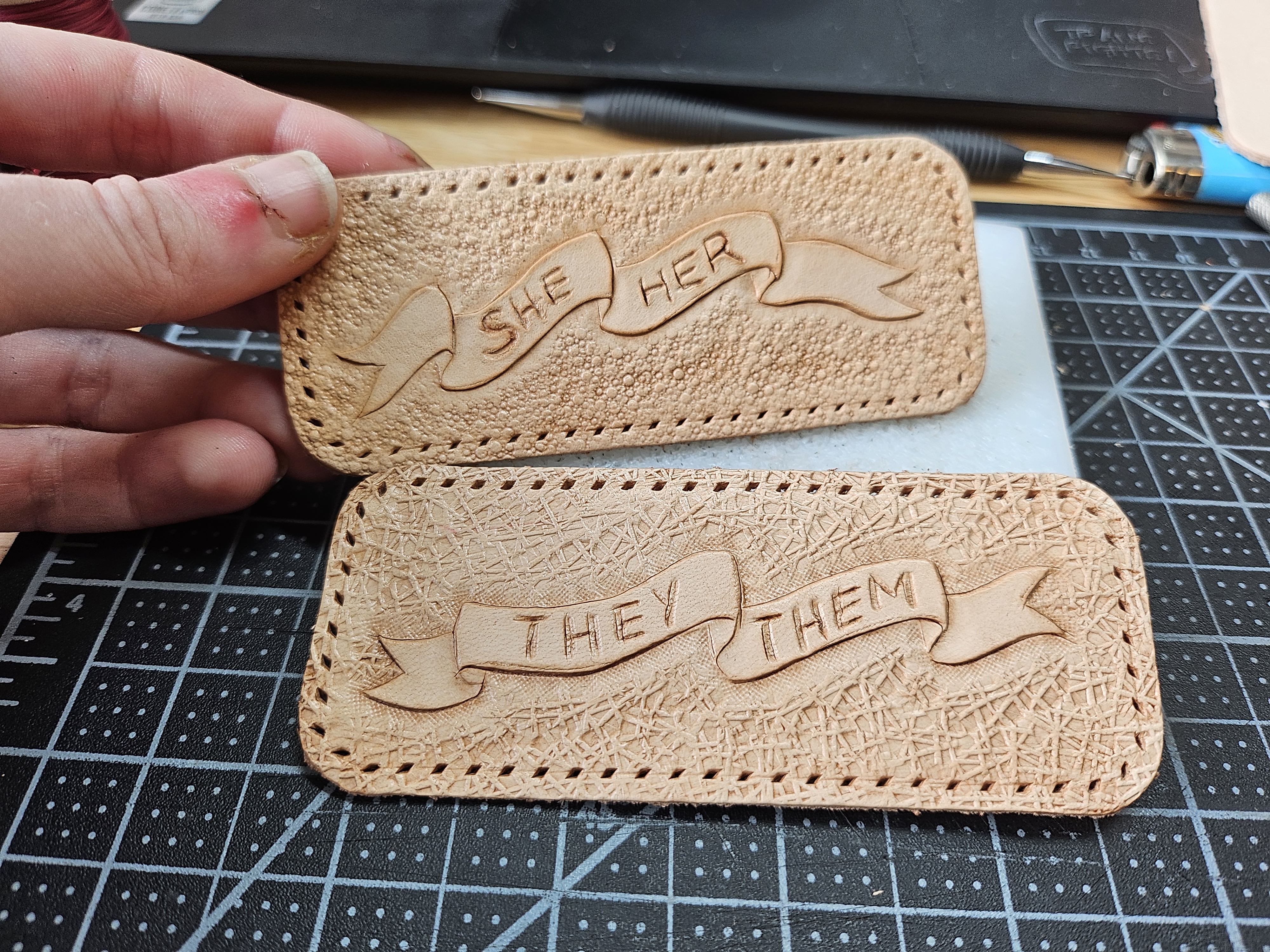 two tooled leather patches. they have scrolls tooled on them that read various pronoun sets.