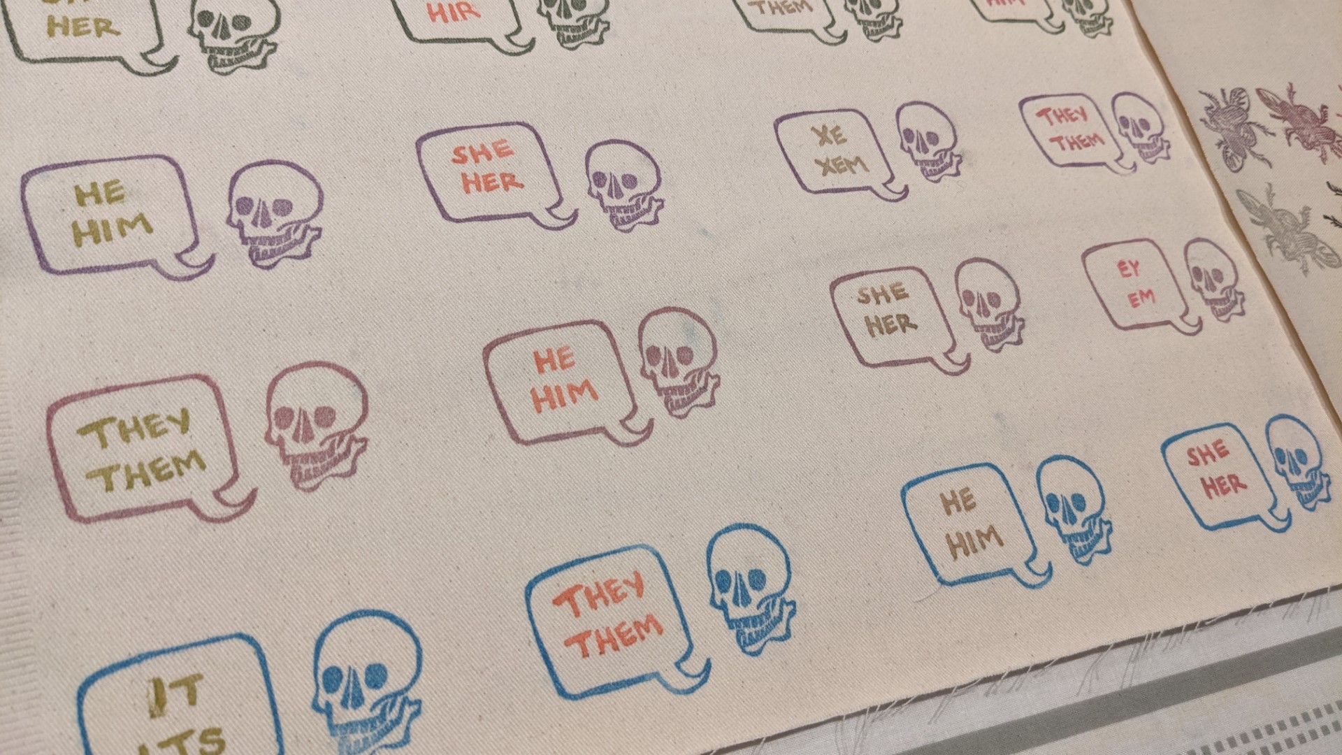 Rows of the same design, a skull with a speech bubble announcing varied pronoun sets, repeat in multiple colors along a stretch of off-white fabric.