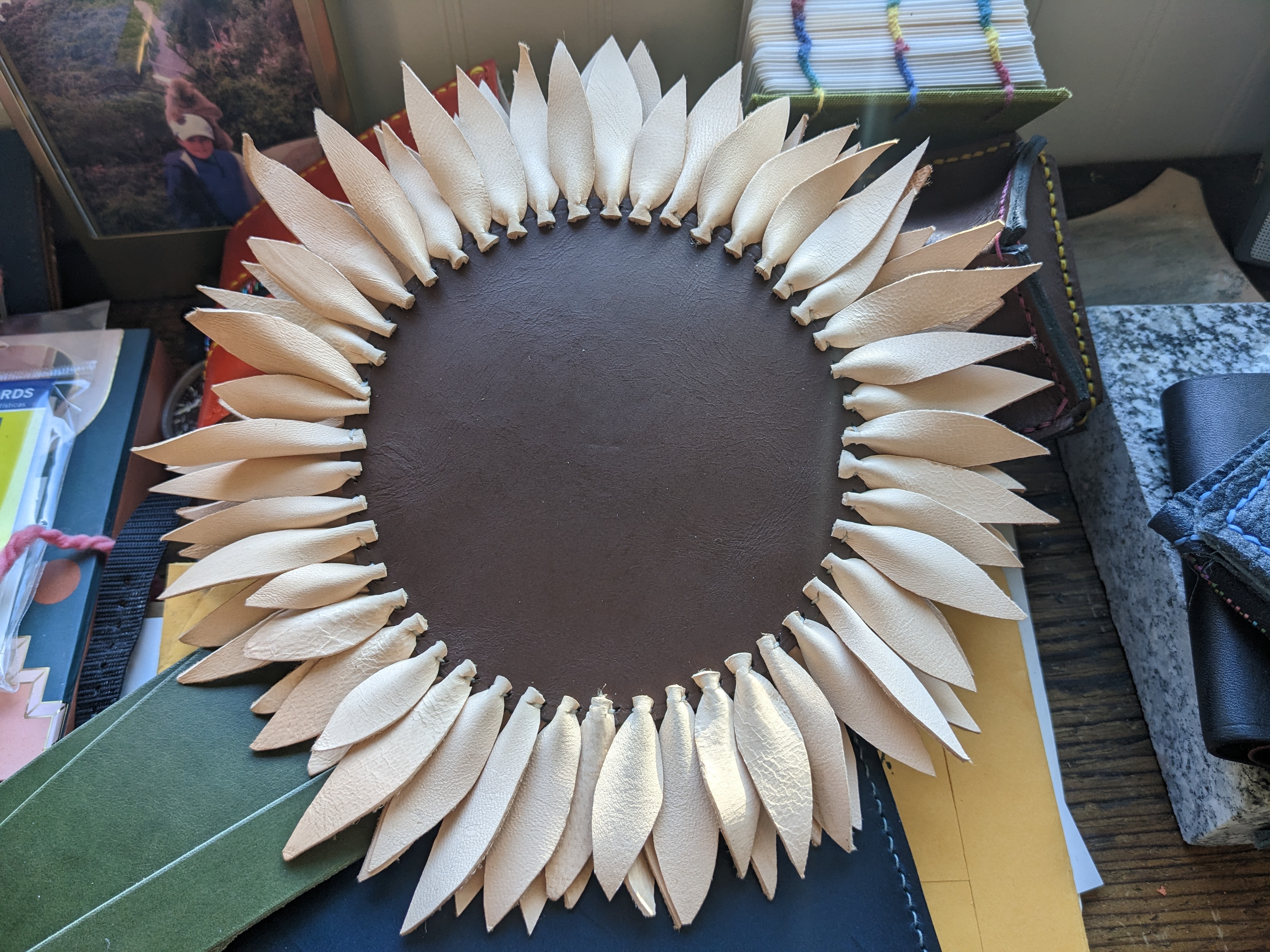 A sunflower made of leather. Many individual natural toned leather petals are sewn onto a brown center .