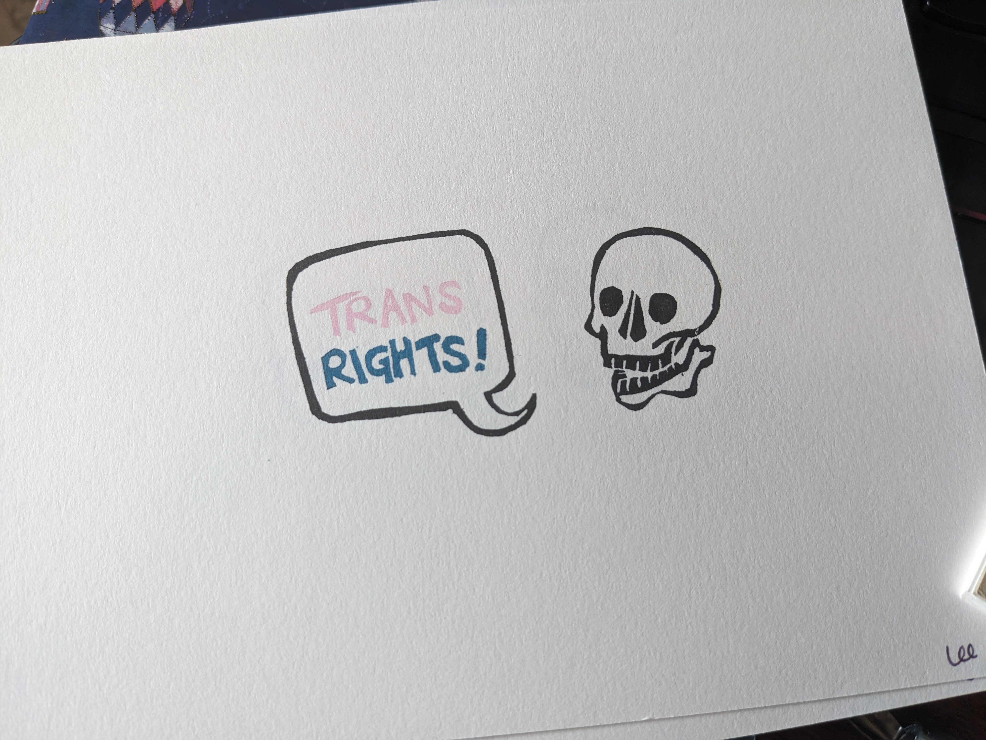 A print in mostly black ink of a smiling skull with a speech bubble. In pink and blue, the speech bubble reads 'trans rights!'