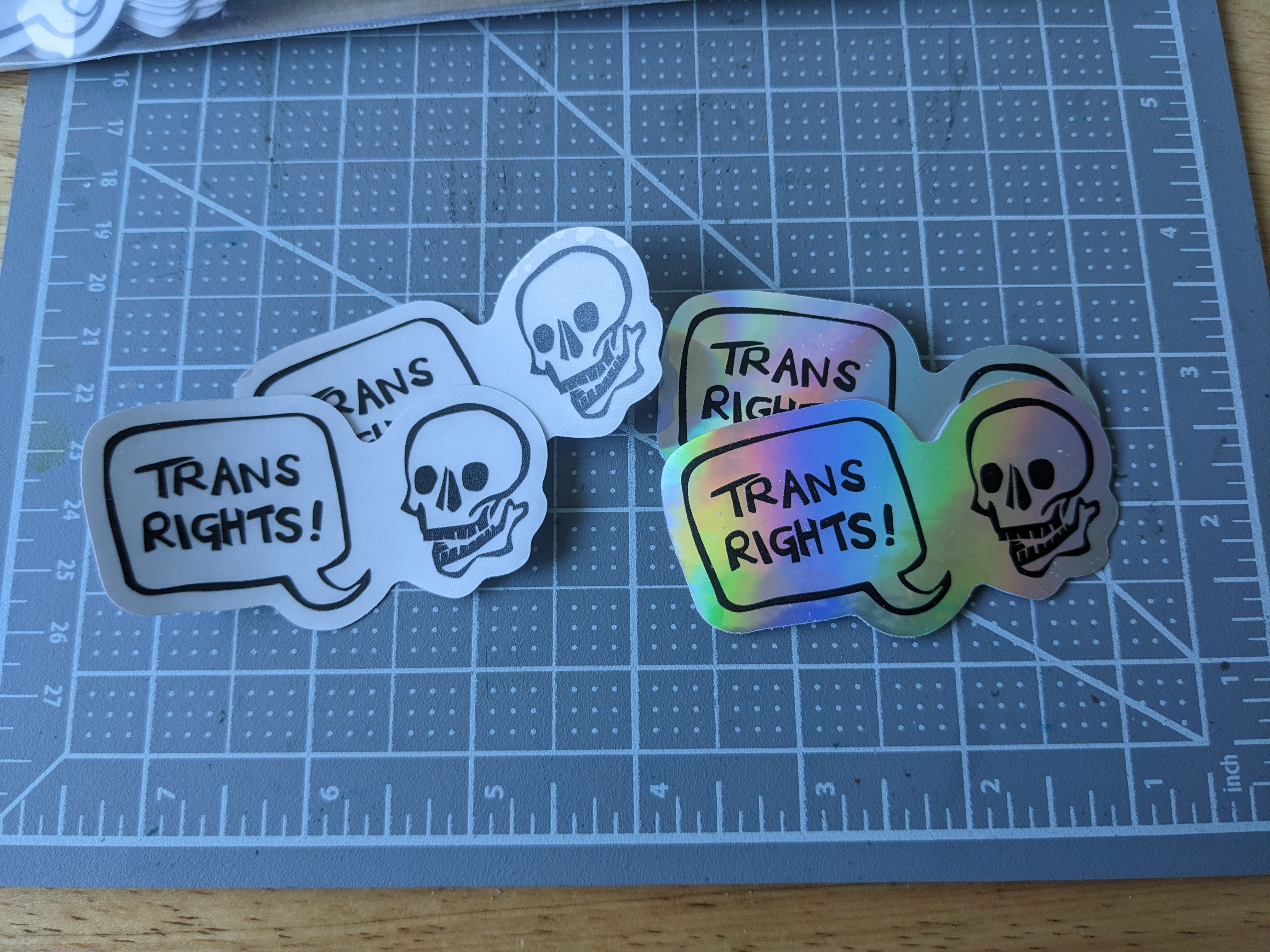 Clear and holographic stickers in the same design - a smiling skull with speech bubble reading 'trans rights!'