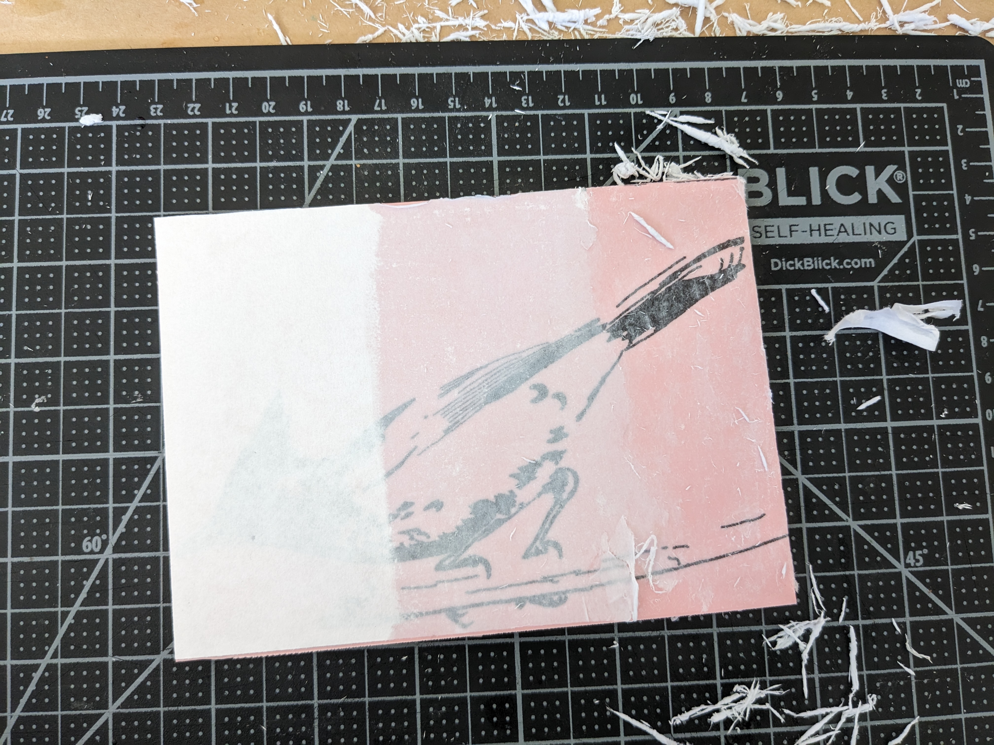 A pink block of carving material with a printed design of a bird attached to it. The paper has been partially rubbed away (process described in this post) to reveal the inked design on the carving material.