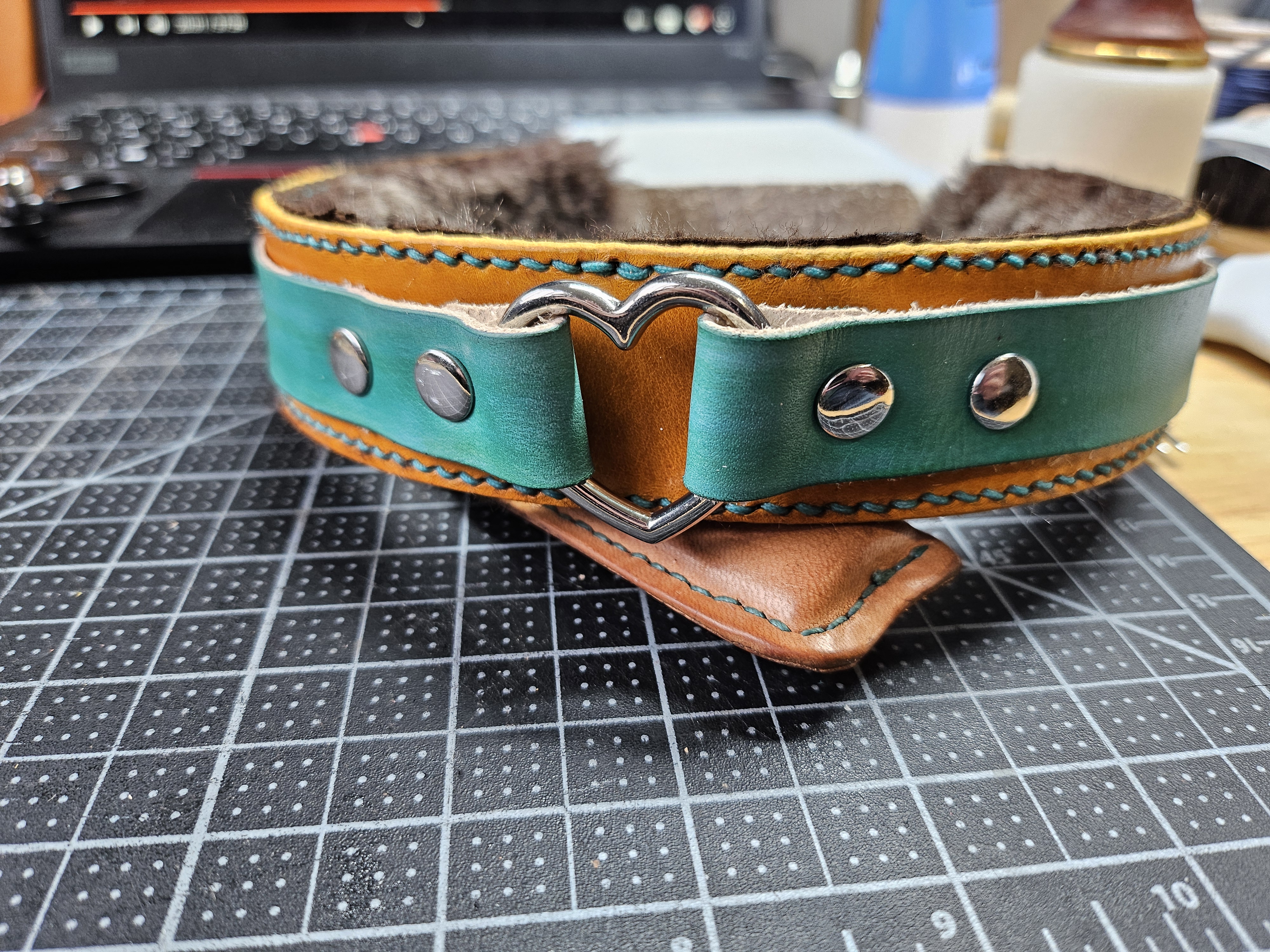A collar rests on a leather-wrapped lighter. It is lined with shearling and built of two other layers of leather - a wider mustard yellow layer and a thinner teal layer over that. the teal layer holds a heart shaped o-ring in place.