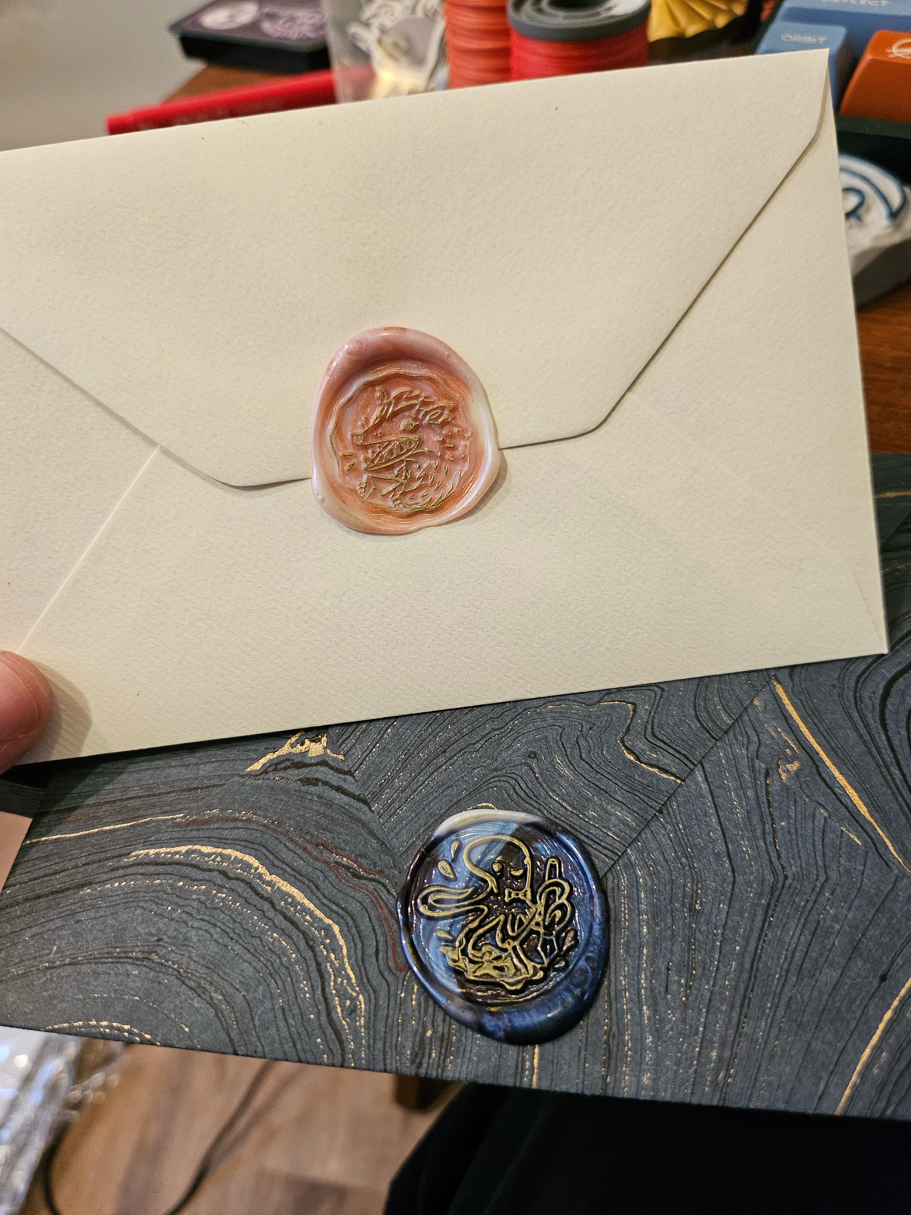 Two envelopes with wax seals. The top envelope is cream with a copper-and-white swirled seal depicting a yelling possum holding mail. The bottom envelope is marbled blue, black, and gold, with a white-and-blue swirled seal depicting an octopus holding a fountain pen.