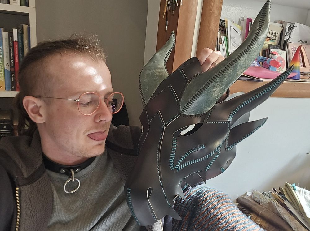 lee (a white person with glasses and a side shave) holds up a leather dragon mask in black and dark green. ze sticks hir tongue out at it.