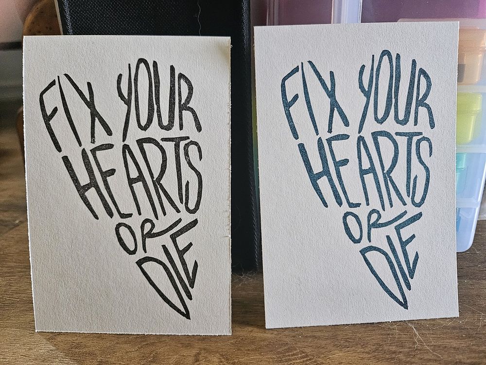2 copies of the same print, one in black ink and one in dark teal. The print is text that reads 'fix your hearts or die', with the text shaped into a somewhat long and narrow heart.