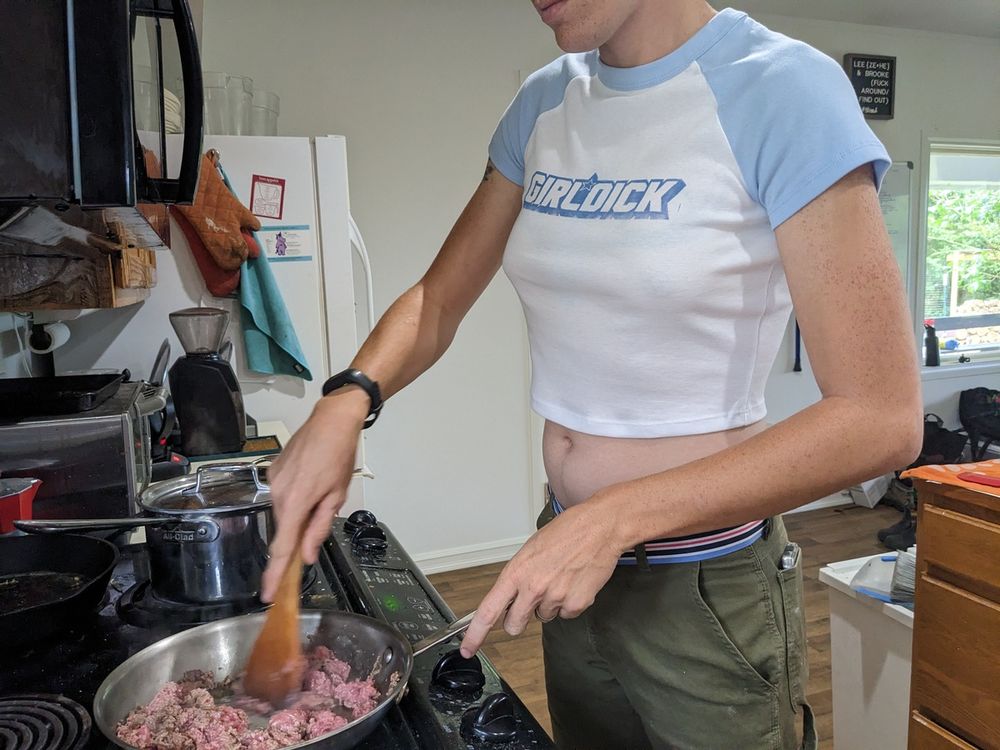 A butch cooking and wearing a cropped tee with blue cap sleeves that reads girldick in blue G.I.Joe font.