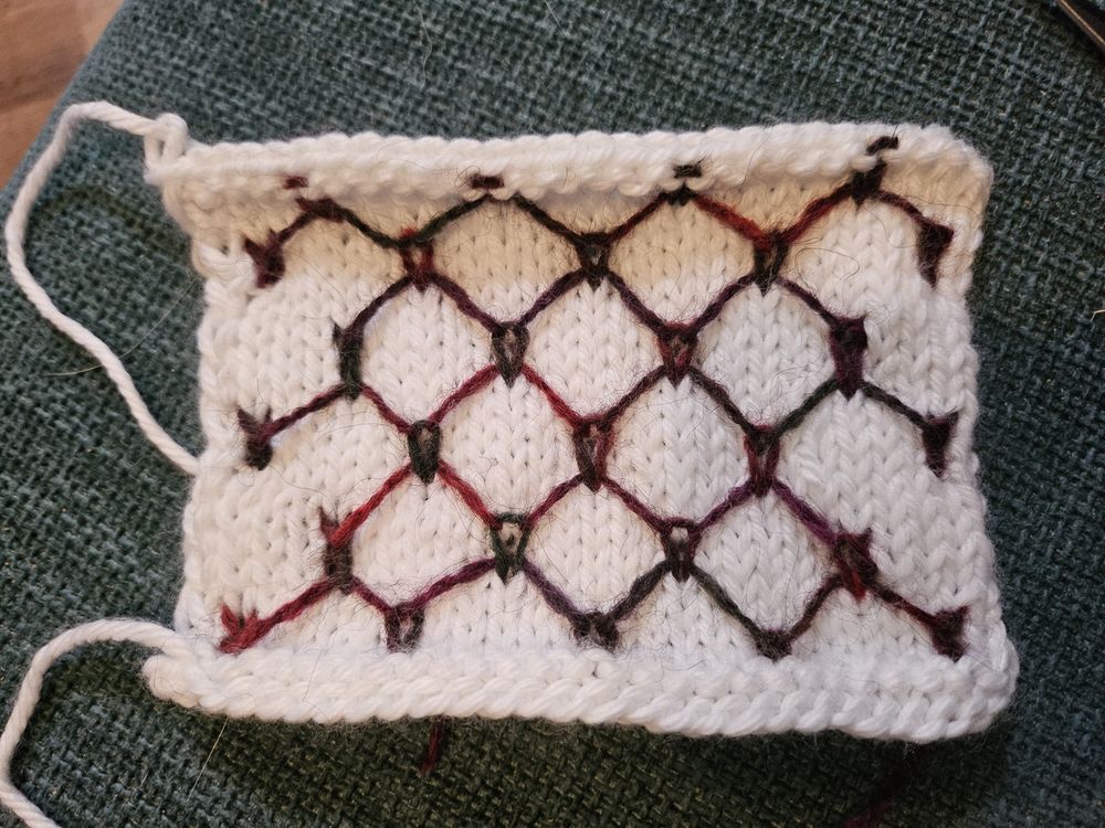 a knitted swatch. it's mostly white yarn, but a grid of dark red- purple- green yarn that looks like a chain link fence is built into it. the diagonal lines are achieved with floats of yarn from one row, then picked up and knit into several rows later.