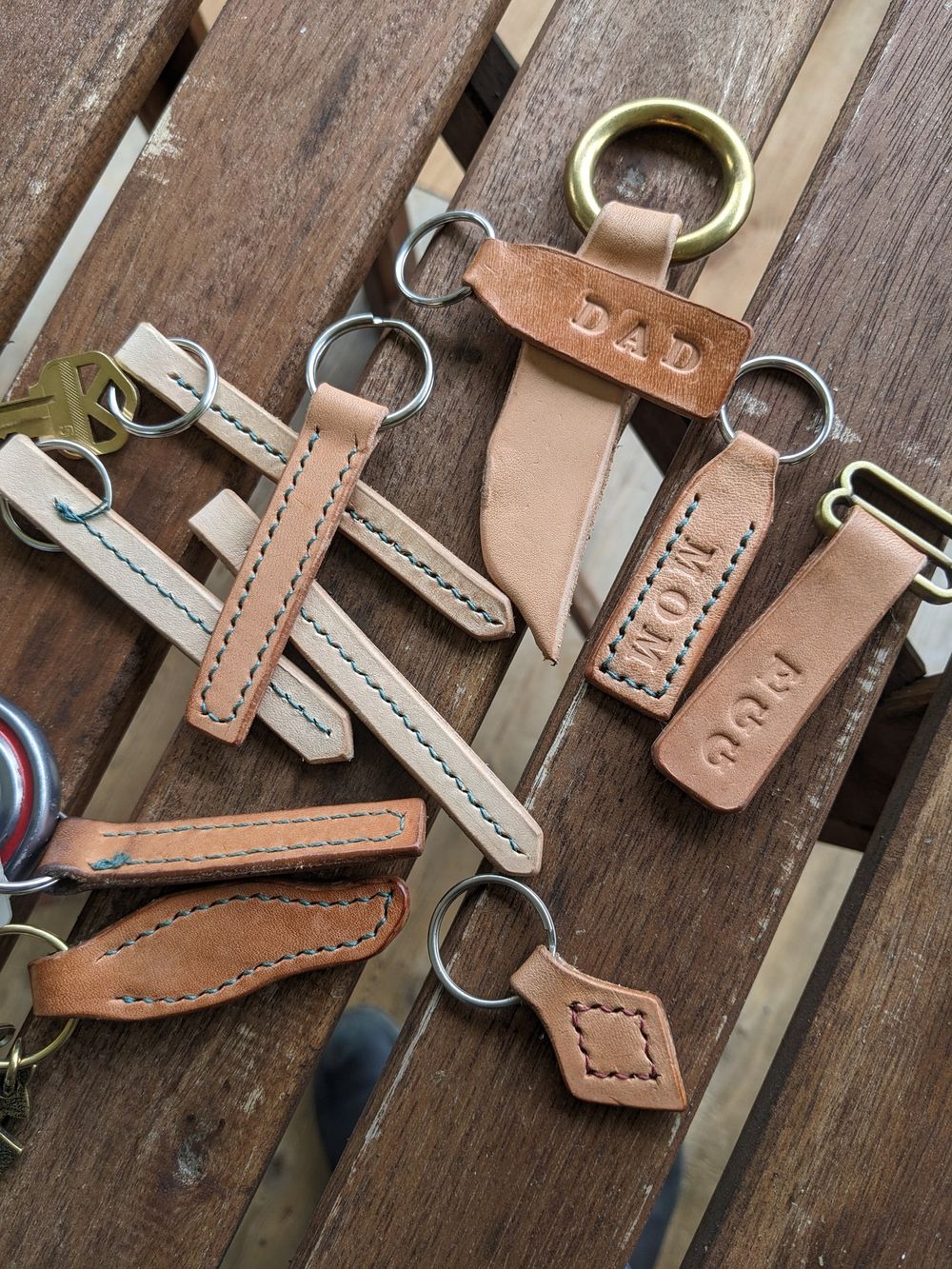 A picture of multiple leather keychains sitting on a wood table. Many of them are simple rectangle shapes with stitching around the edge; a few are odd wavy or geometric shapes. A few say things like 'MOM' or 'EGG'.