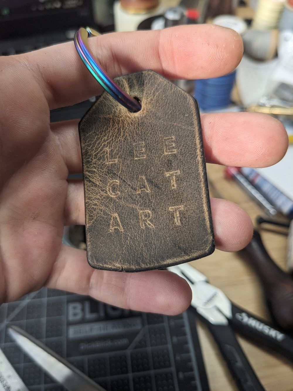 A keychain on an iridescent rainbow split ring. It is dark brown/grey leather and has LEE CAT ART stamped into it.
