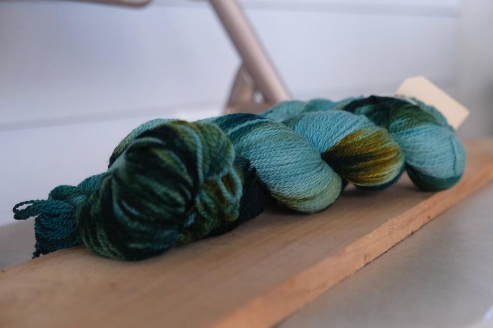 1 large and 2 small skeins of handspun yarn in a sport or DK weight that has been dyed in light and dark teal, green, and rusty green-gold.