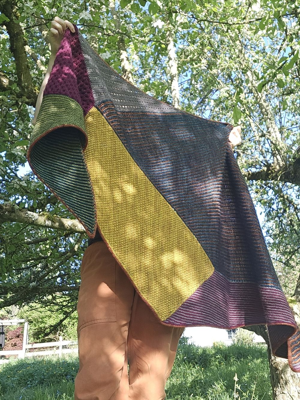 a largely unseen person holds up an expansive knitted shawl, built in 6 sections of different textures and colors. in the background, trees and dappled sunlight.